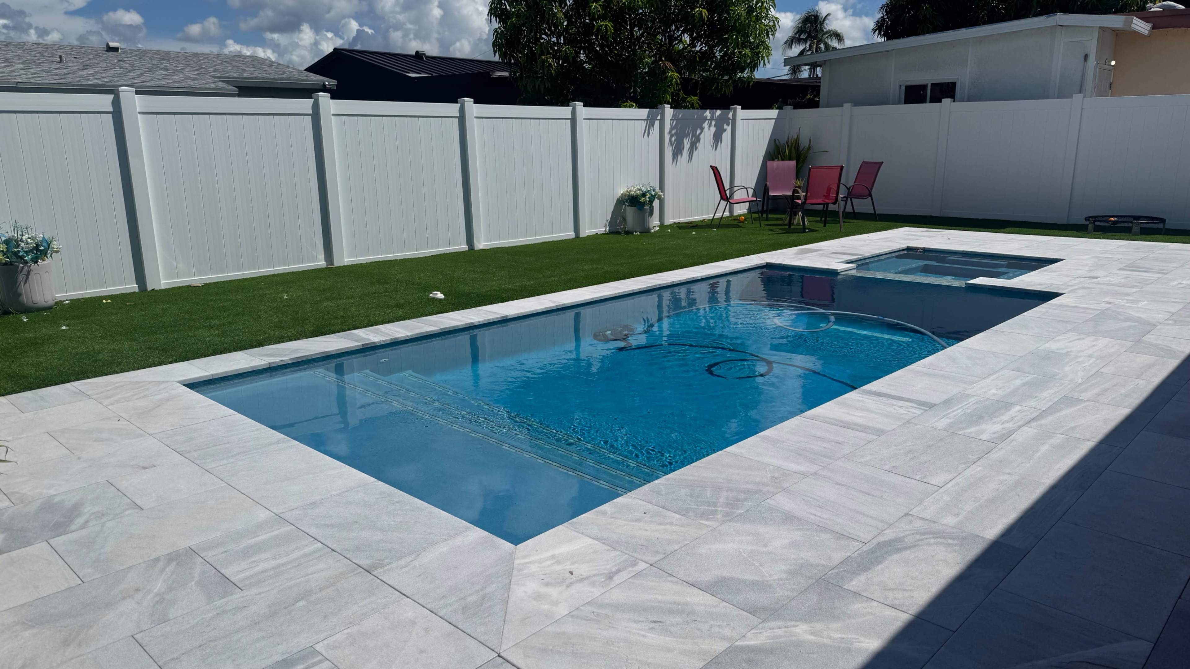 The image shows a rectangular swimming pool with a spa, surrounded by a patio area and a white fence, set against a backdrop of blue skies and scattered clouds.