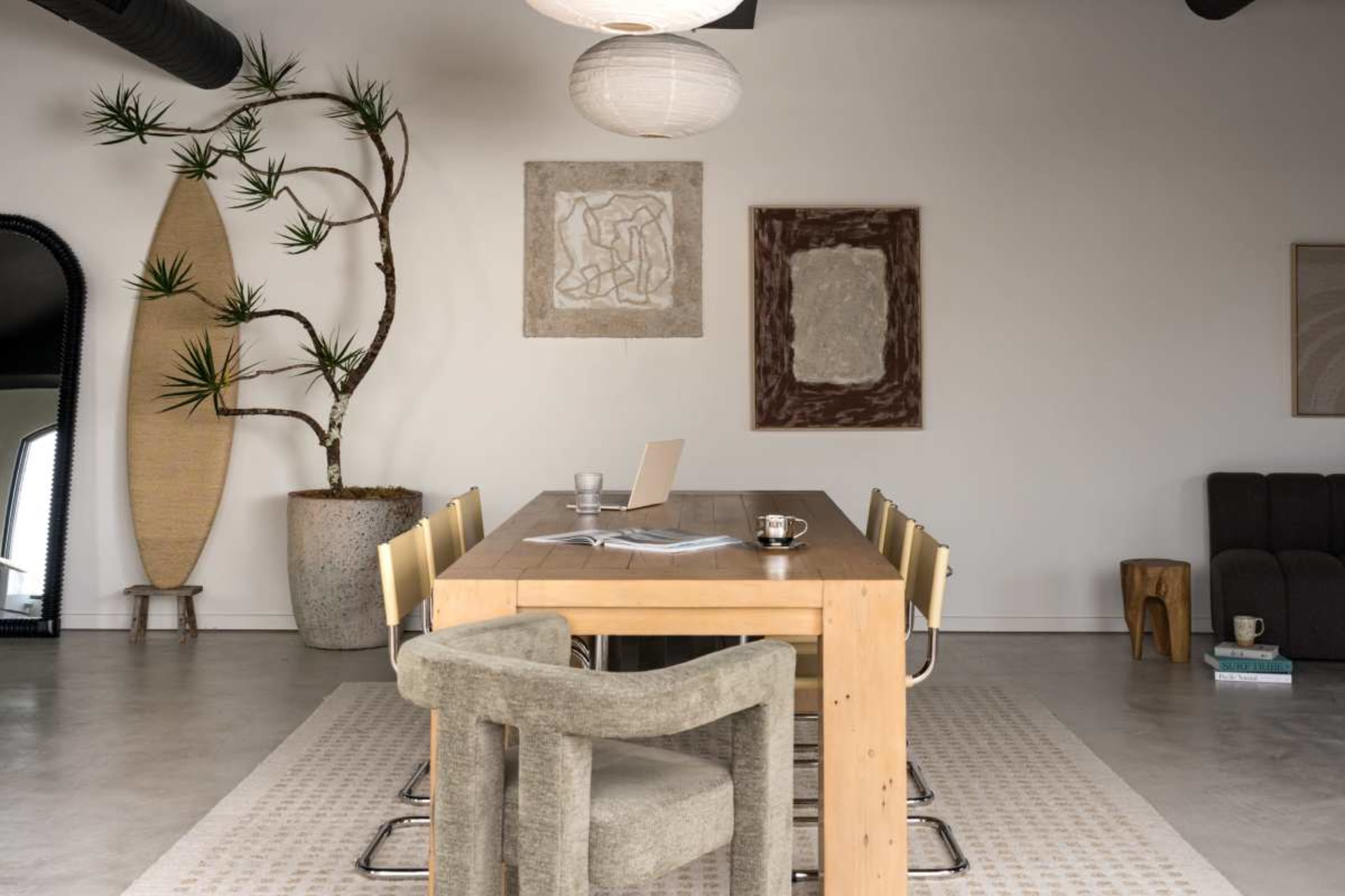 A spacious dining area features a long wooden table surrounded by chairs, with abstract art and a large indoor plant on the walls.
