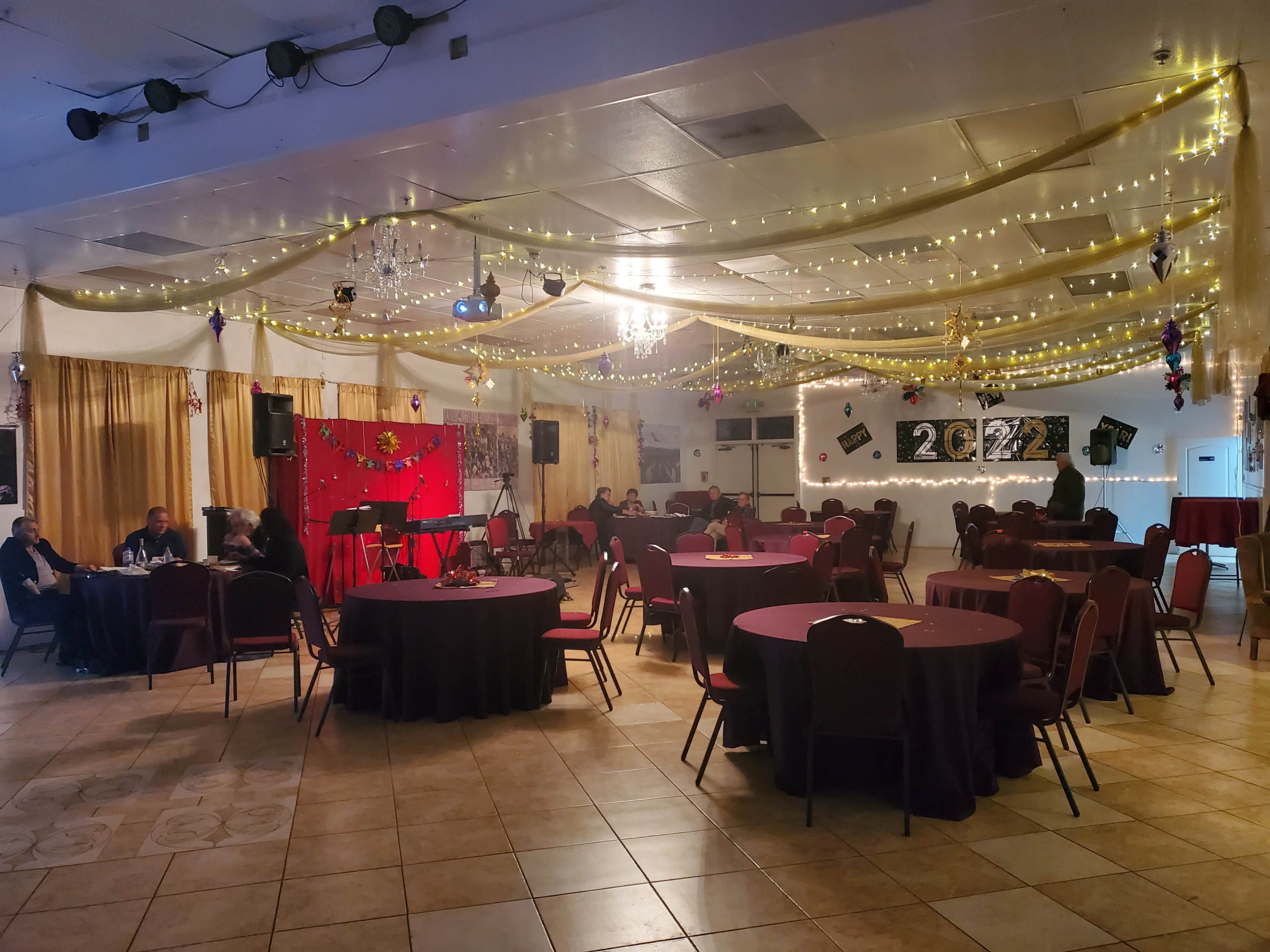 A banquet hall decorated with string lights and a stage set up for a performance, featuring round tables and chairs arranged for an event.