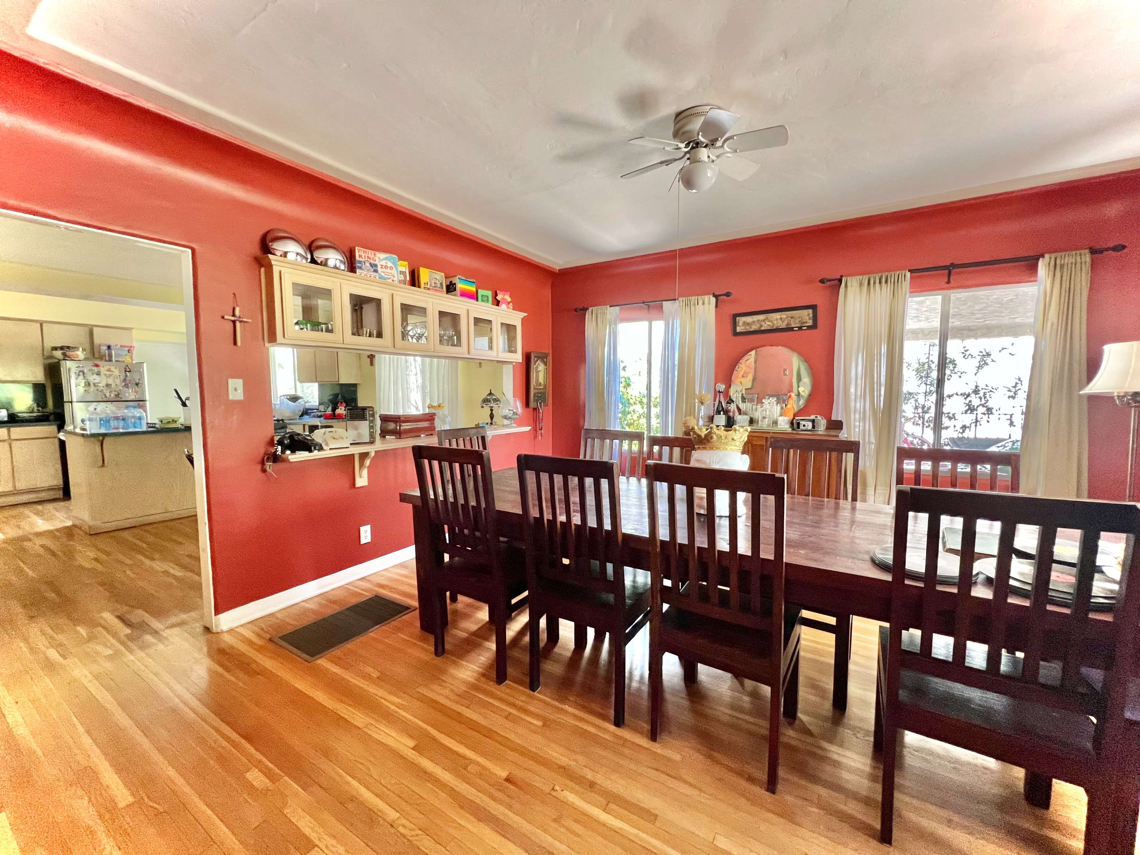 A dining room features a large wooden table surrounded by eight chairs, with red walls and a window providing natural light.