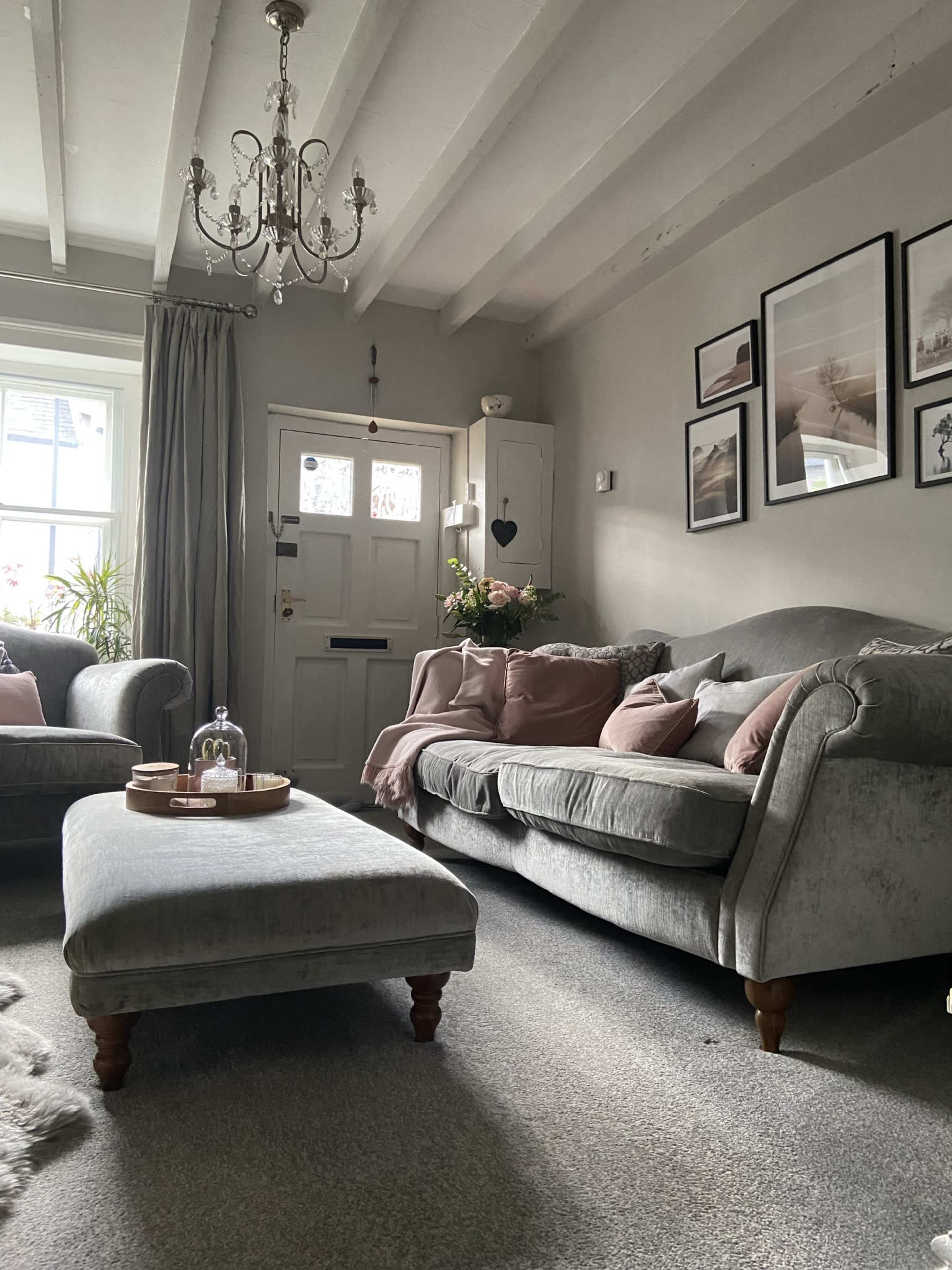The living room features two gray sofas with pink cushions, a decorative ottoman in the center, and a chandelier hanging from the beamed ceiling.