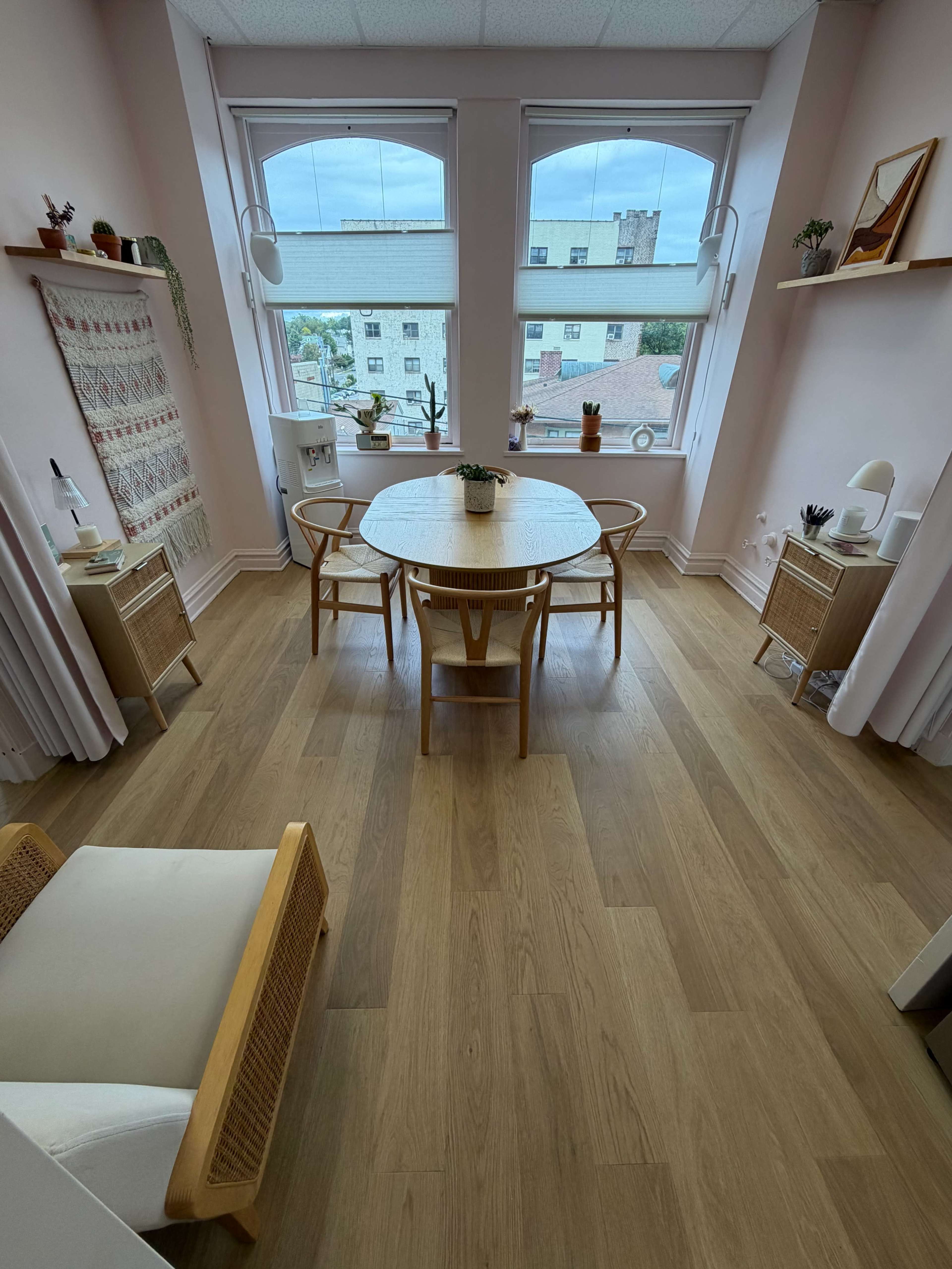 The image shows a light-filled room with a round wooden dining table surrounded by four chairs, complemented by large windows and decorative shelves.