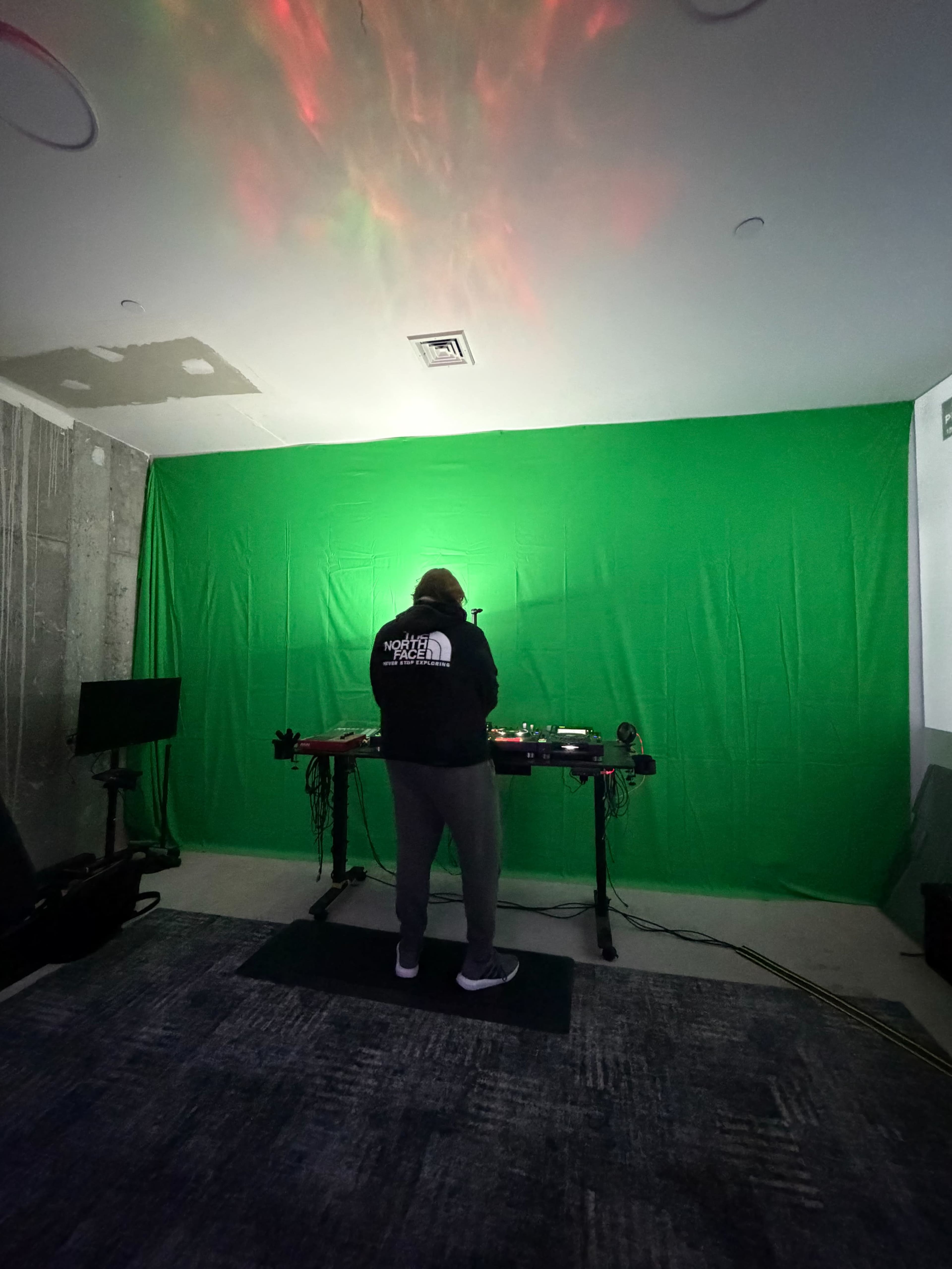 A person stands in front of a green backdrop, with a lighting setup casting colored lights on the scene, while using equipment on a table.