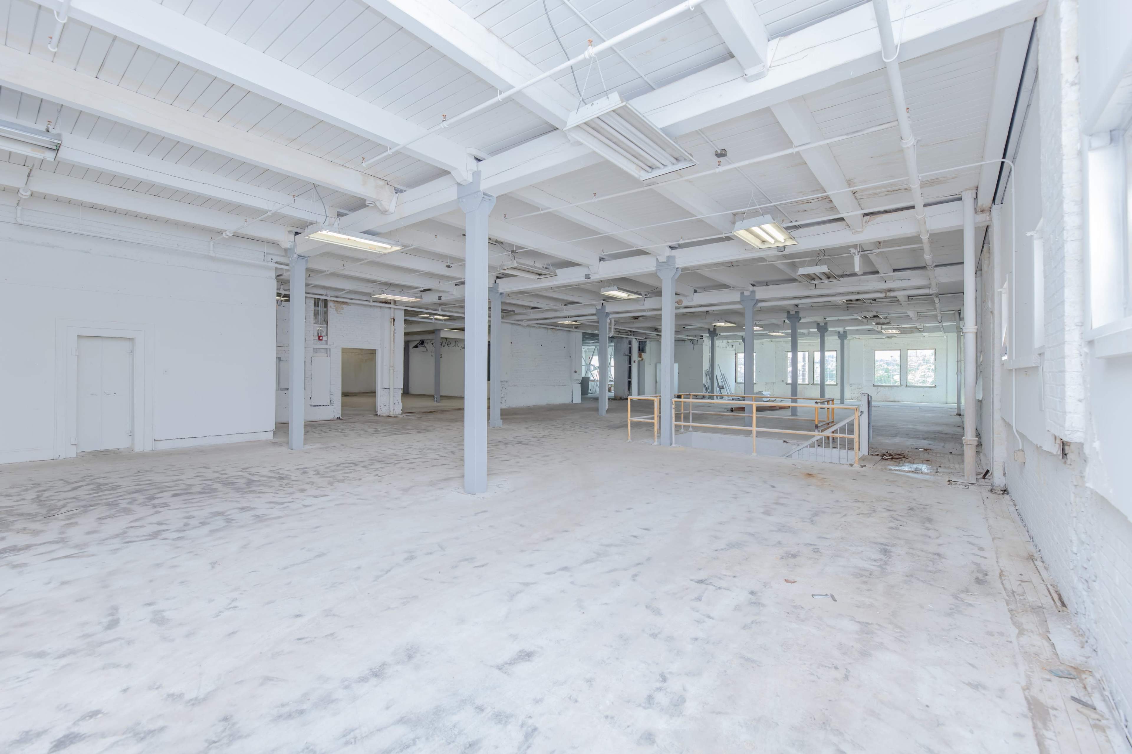 The image shows a spacious, empty industrial room with white walls and a concrete floor, featuring large windows and a railing area in the background.