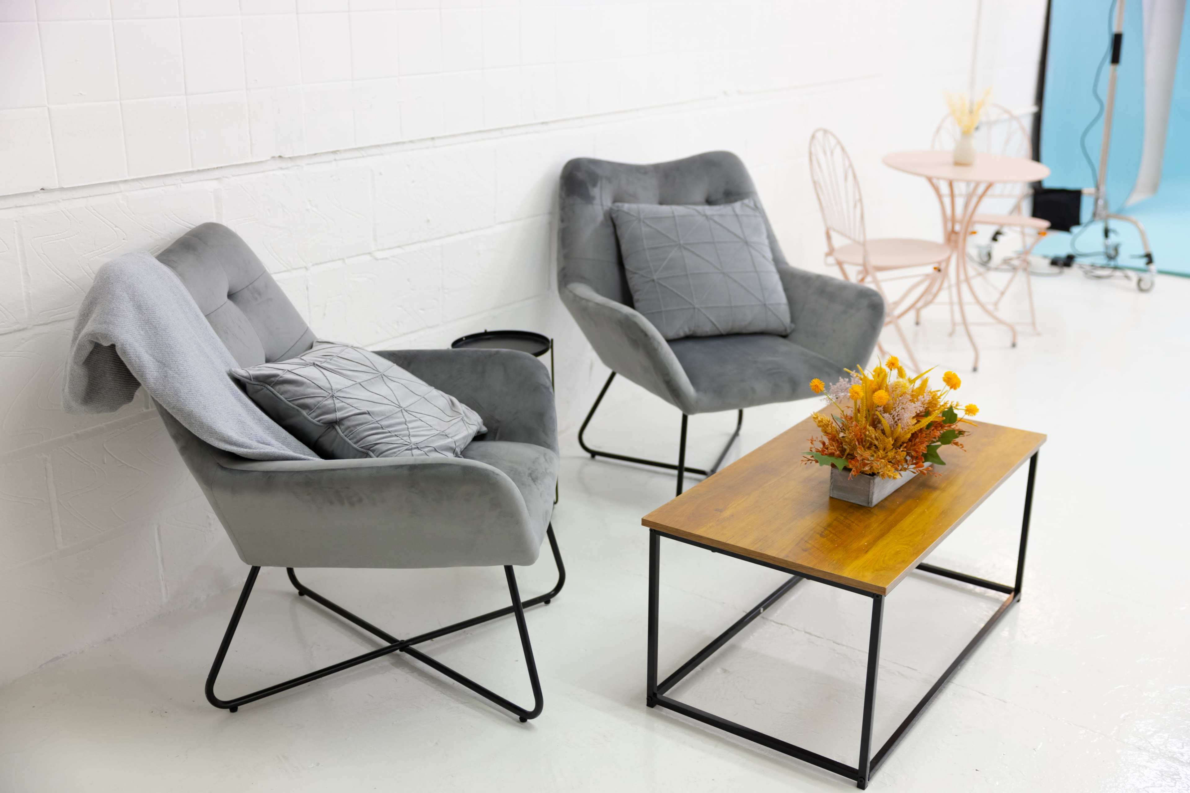 The image shows two grey armchairs with cushions, a wooden coffee table with a flower arrangement, and a light-colored side table in a minimalist interior setting.