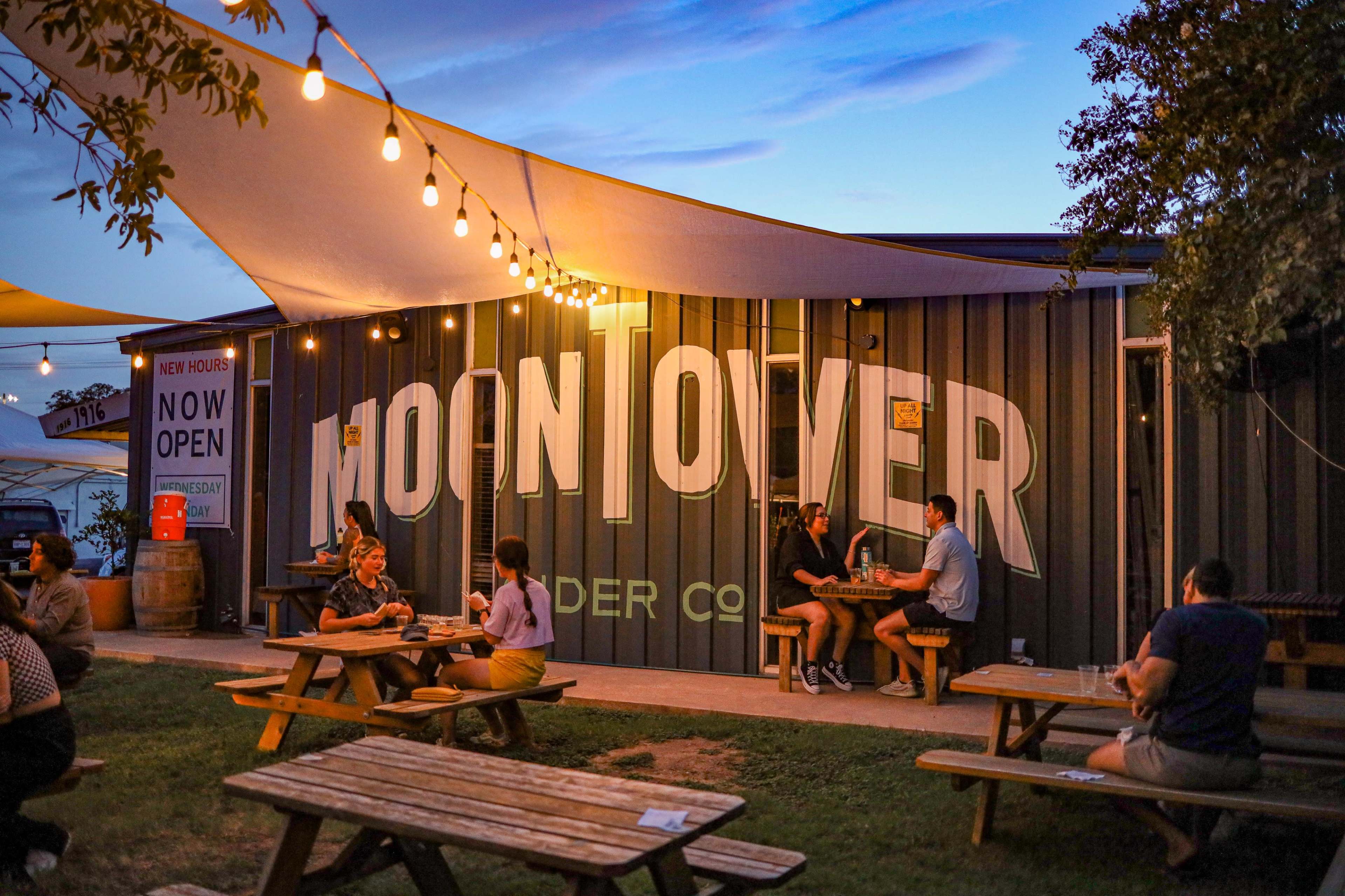 People are seated at picnic tables outside the Moontower Cider Company, enjoying the evening ambiance under string lights.