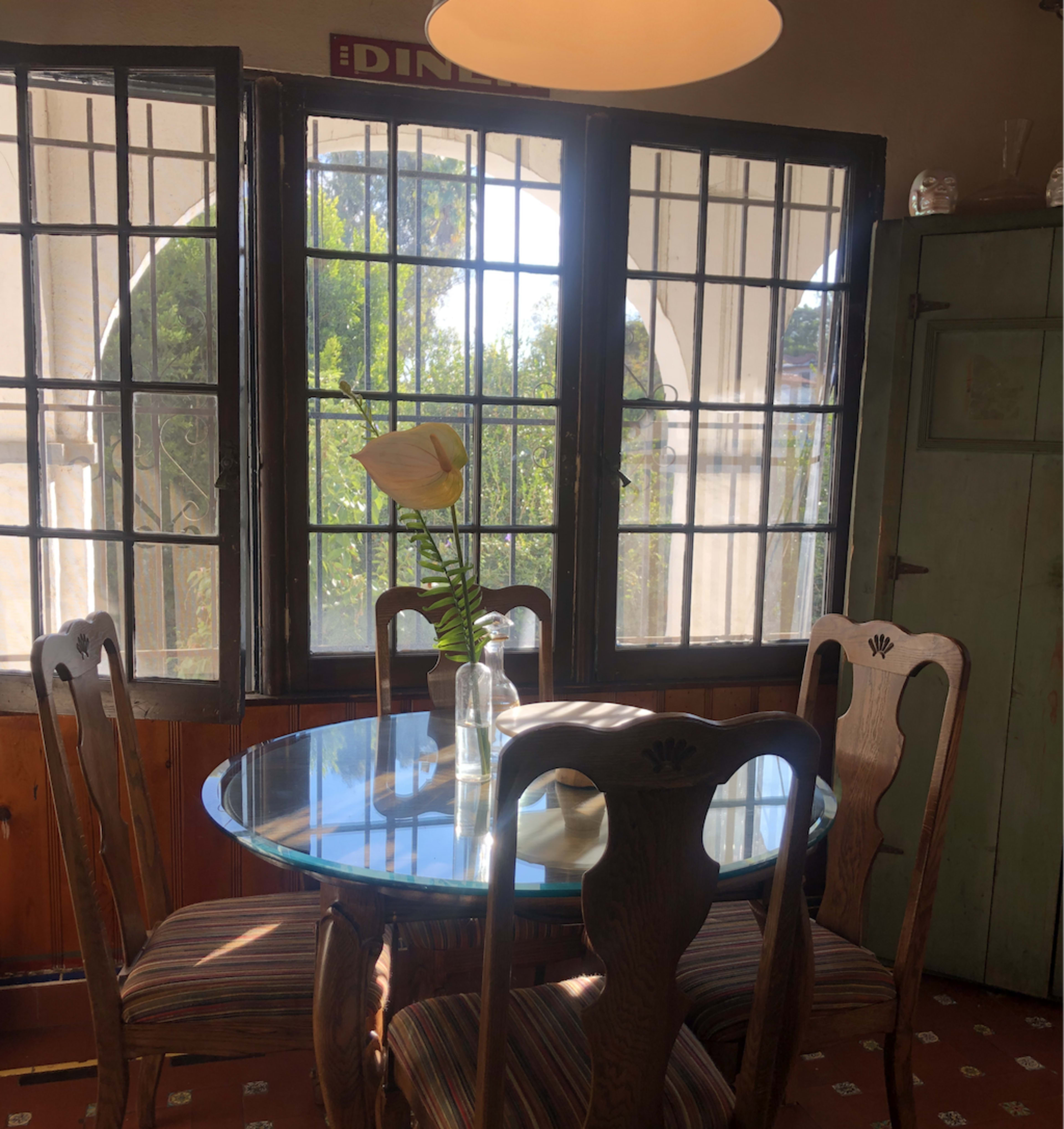 A circular glass dining table is set with four wooden chairs, surrounded by large windows that let in natural light and view greenery outside.