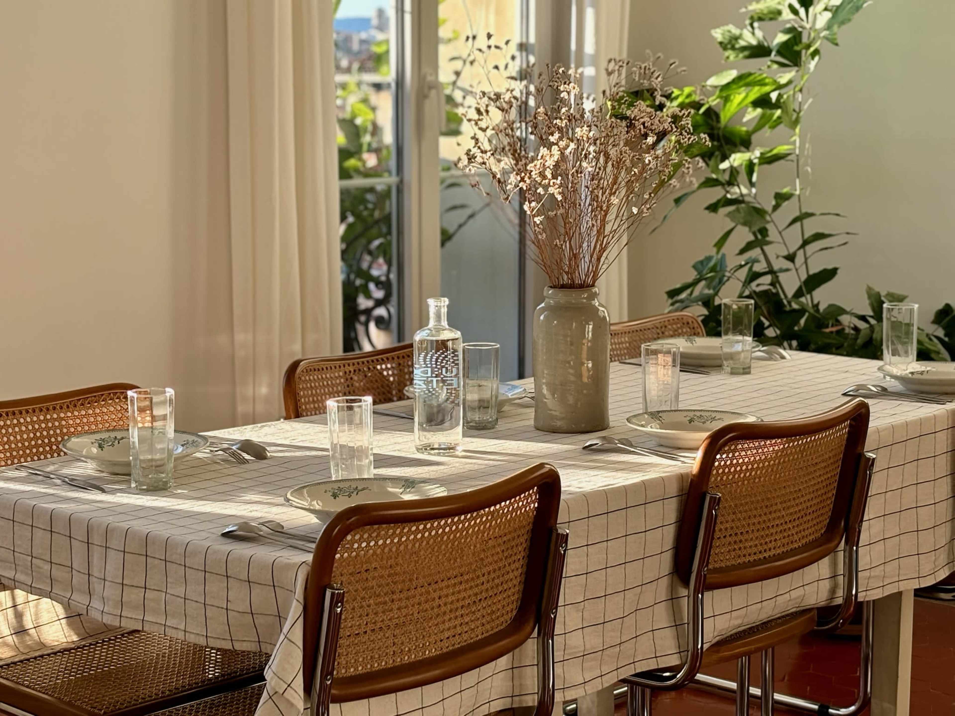 A neatly set dining table with a checkered tablecloth features glassware, plates, and a vase of dried flowers as a centerpiece.