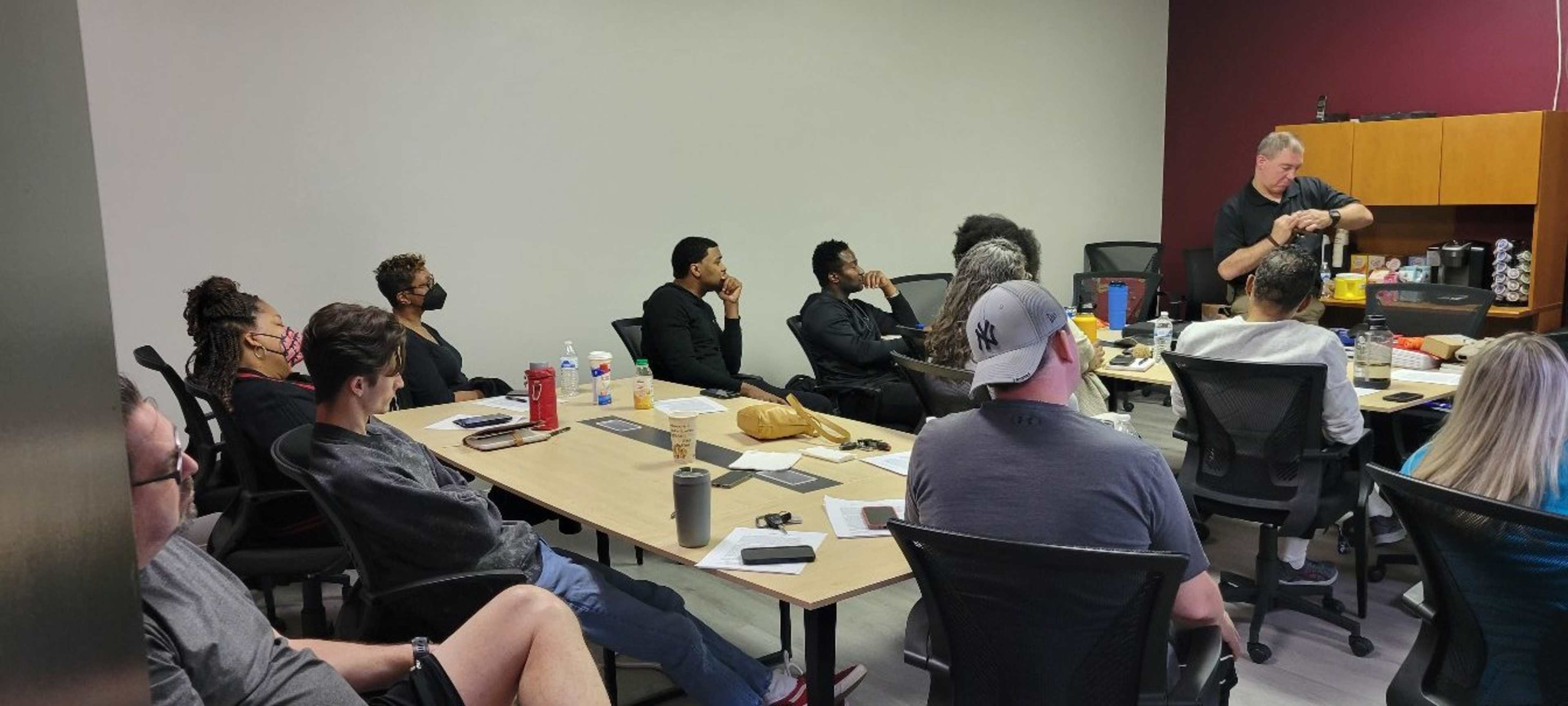 A group of individuals sits in a conference room, attentively listening to a speaker at the front while surrounded by various materials and drinks on the table.