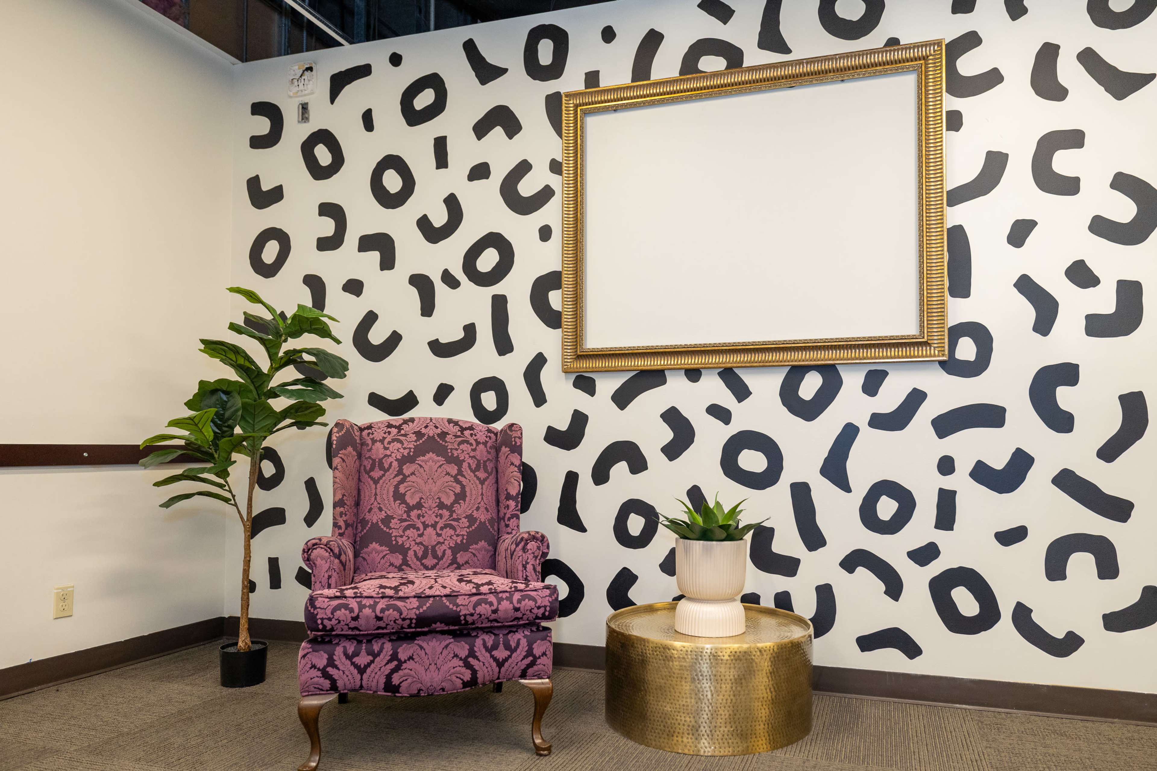 The image shows a corner of a room featuring a purple patterned armchair, a gold circular table, a potted plant, and a wall adorned with black abstract shapes and a large empty picture frame.
