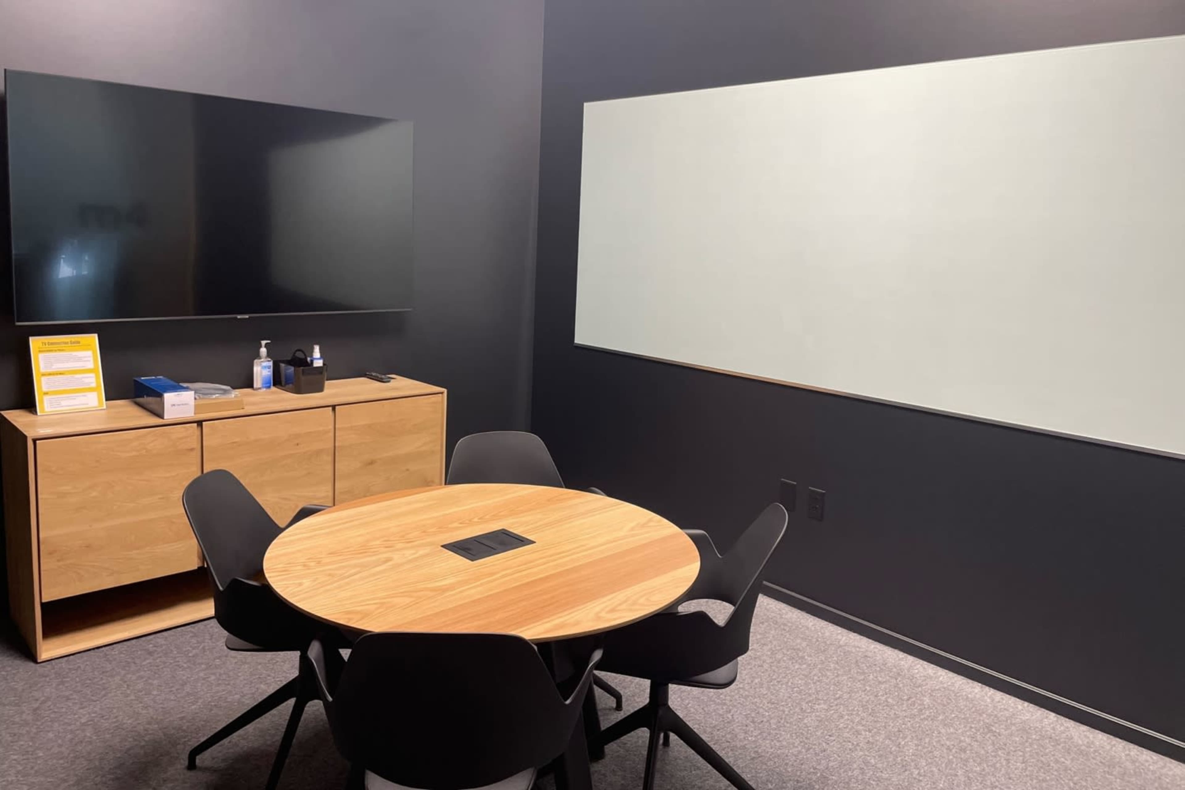 A meeting room features a round wooden table surrounded by black chairs, a wall-mounted flat-screen TV, and a blank whiteboard.