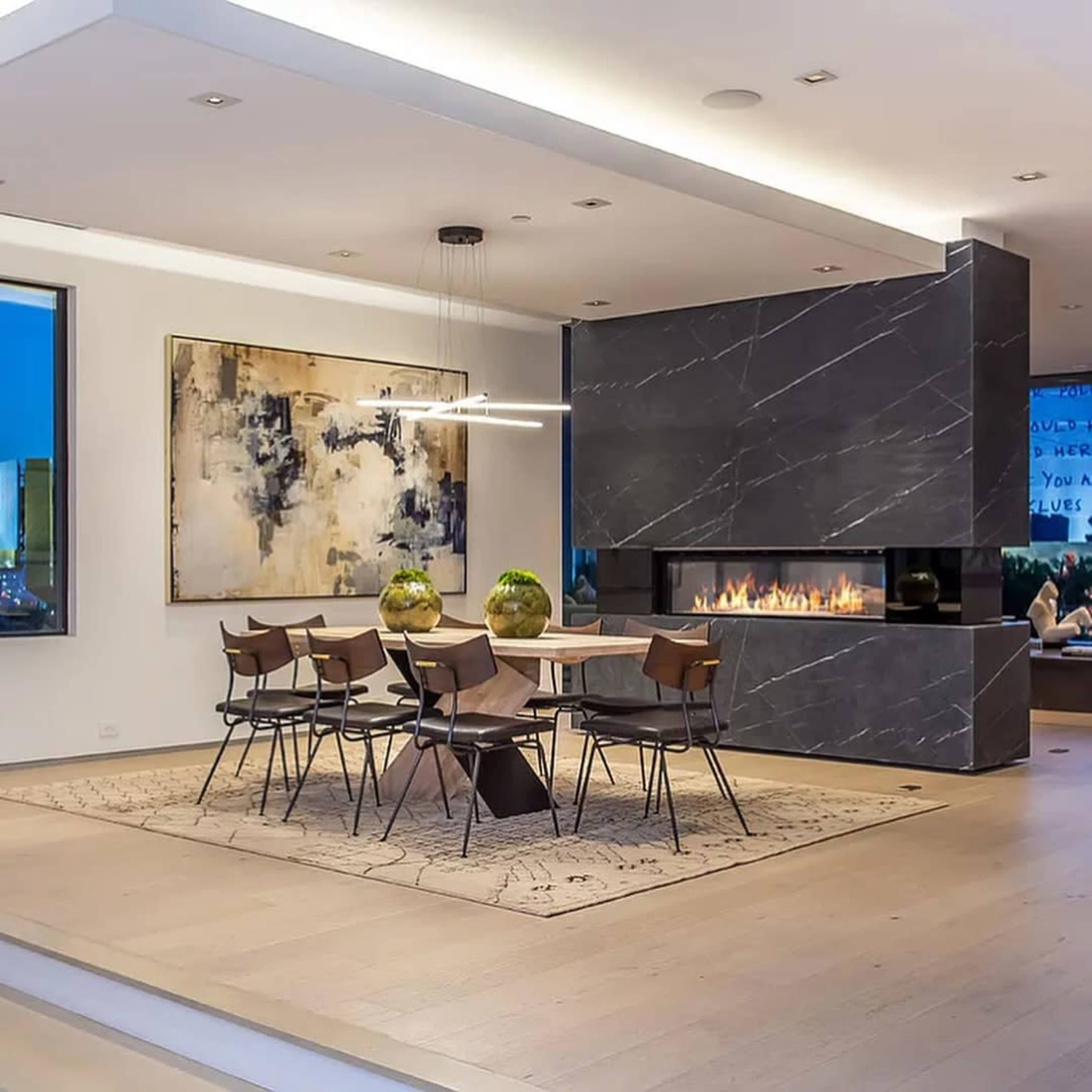 A modern dining area features a stone table surrounded by six chairs, with a large painting and a fireplace integrated into the design.