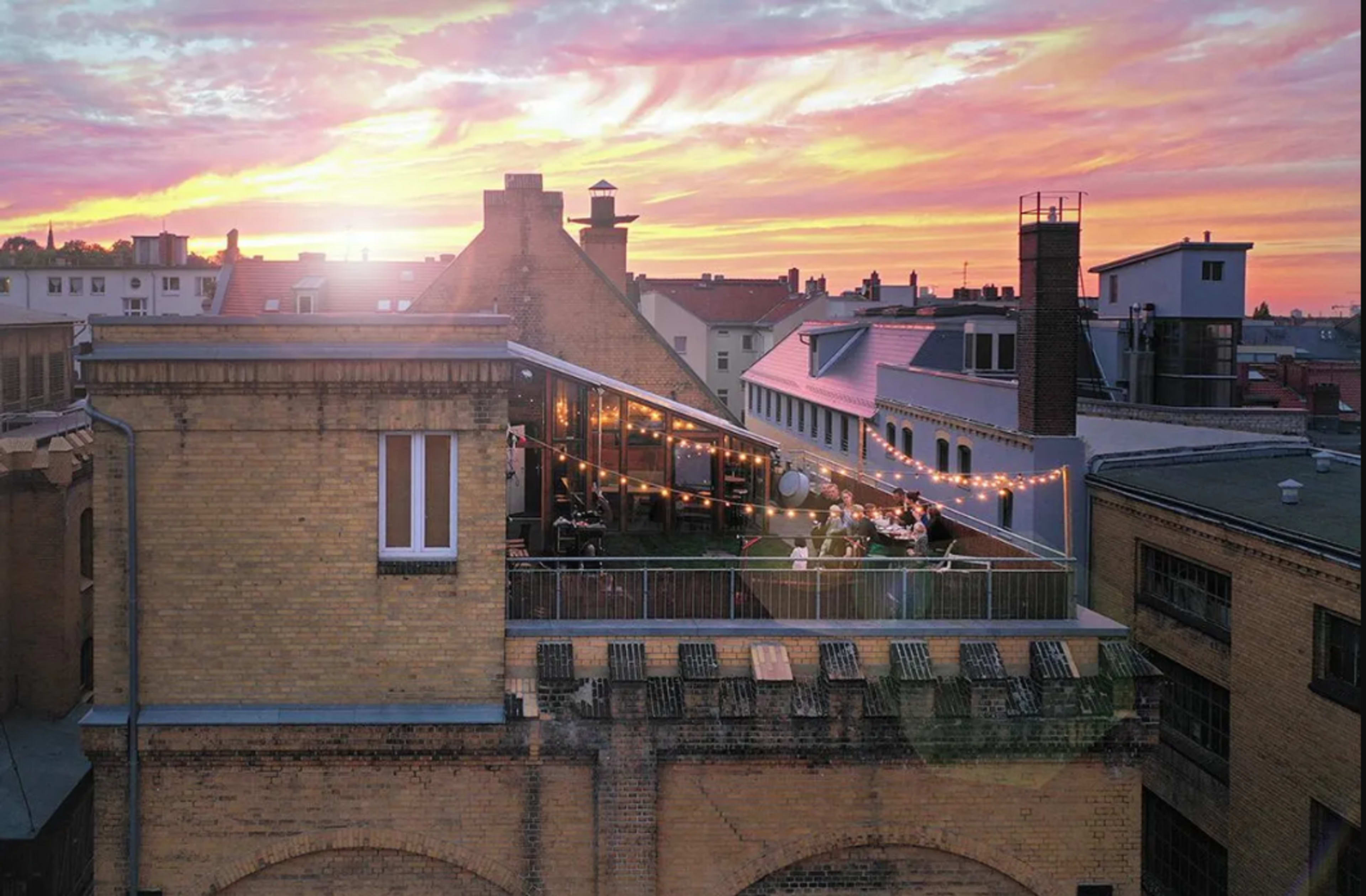 A rooftop gathering is illuminated by string lights against a vibrant sunset sky.