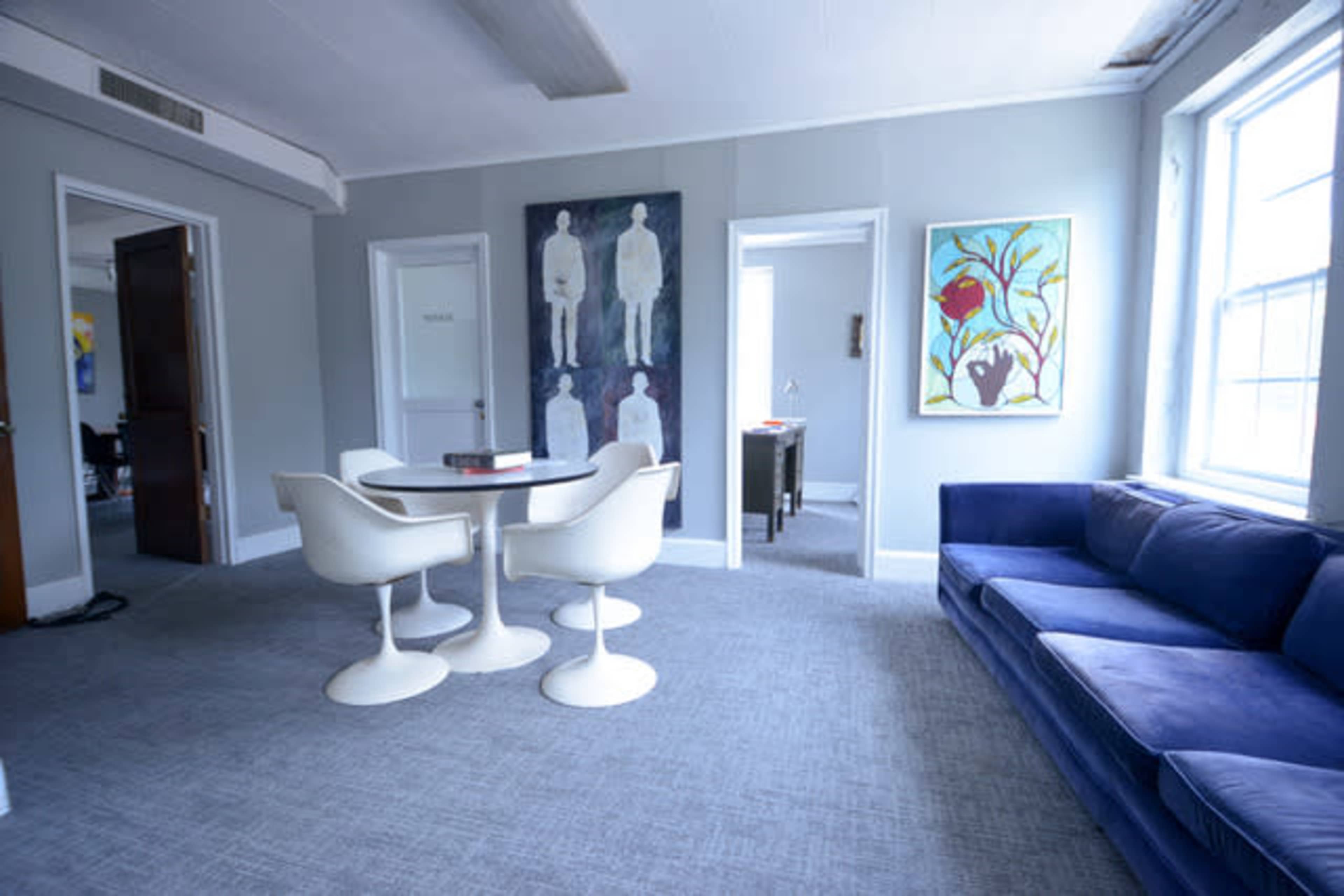 A modern living room featuring a blue sofa, a round dining table with two chairs, and artwork on the walls, with a doorway leading to another room.