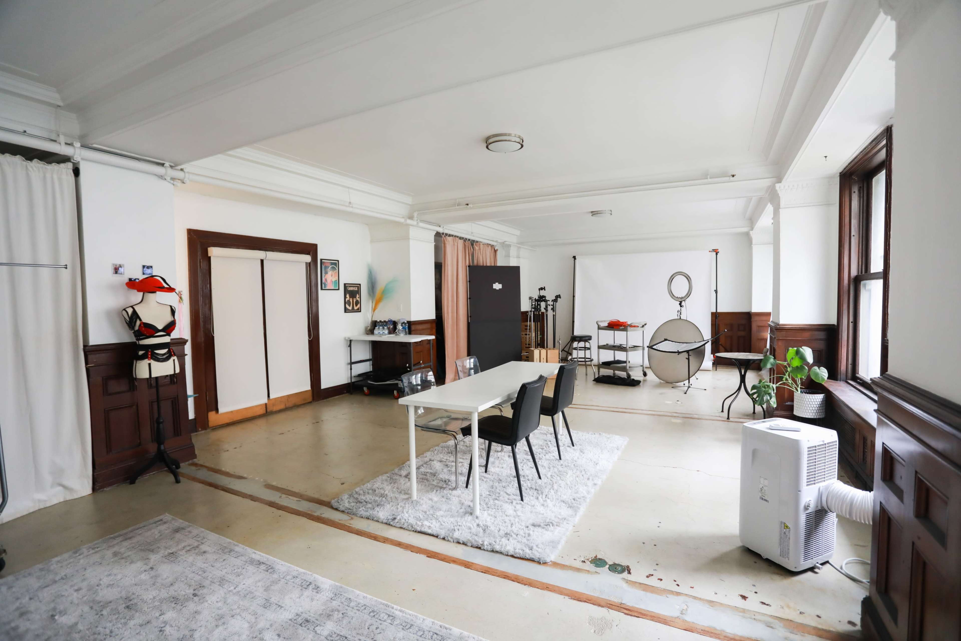The image shows a spacious studio room with a white table and four black chairs, accompanied by photography equipment and a backdrop.