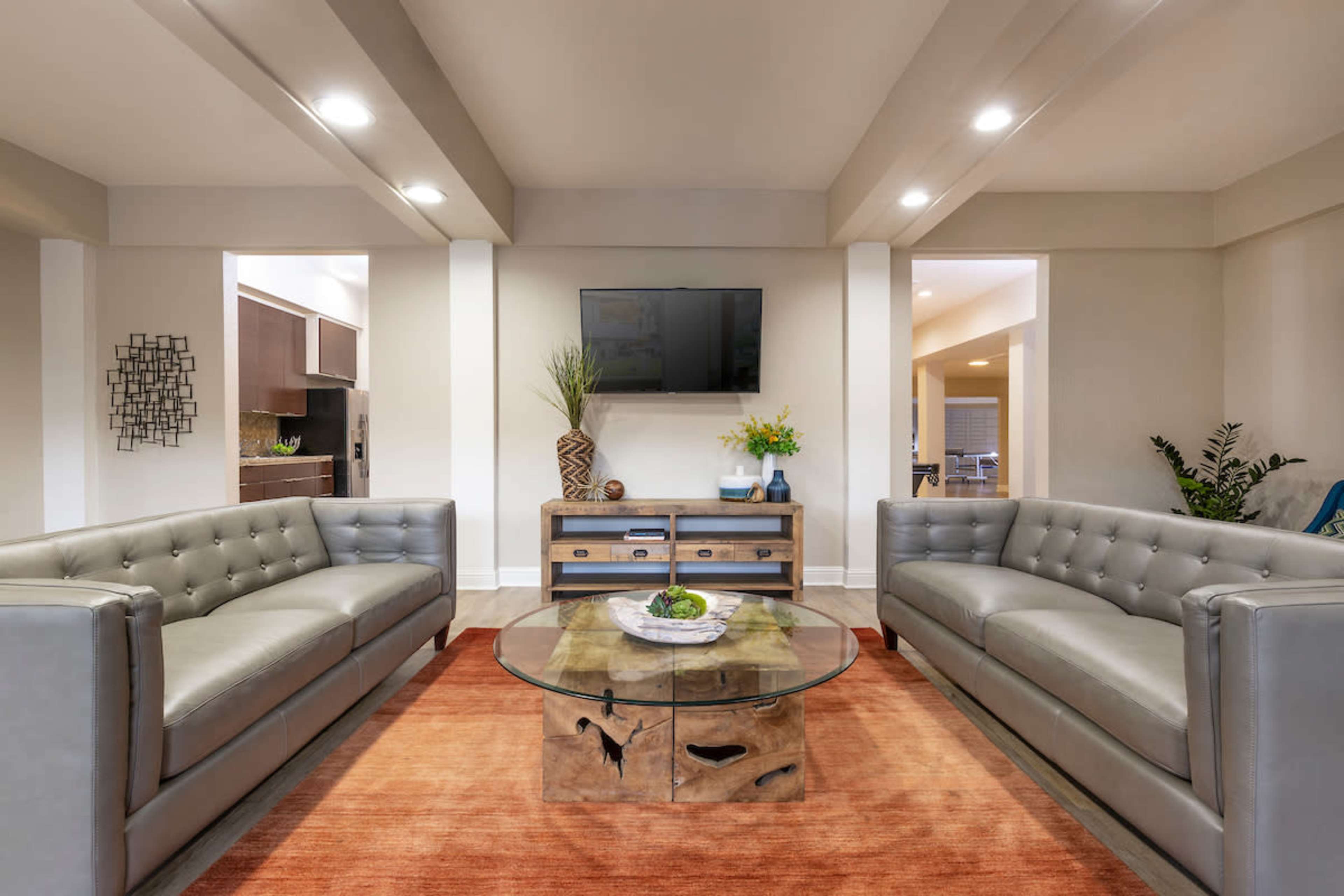 A modern living room featuring two gray sofas facing a wooden coffee table with a glass top, set on an orange area rug, with a television mounted on the wall.