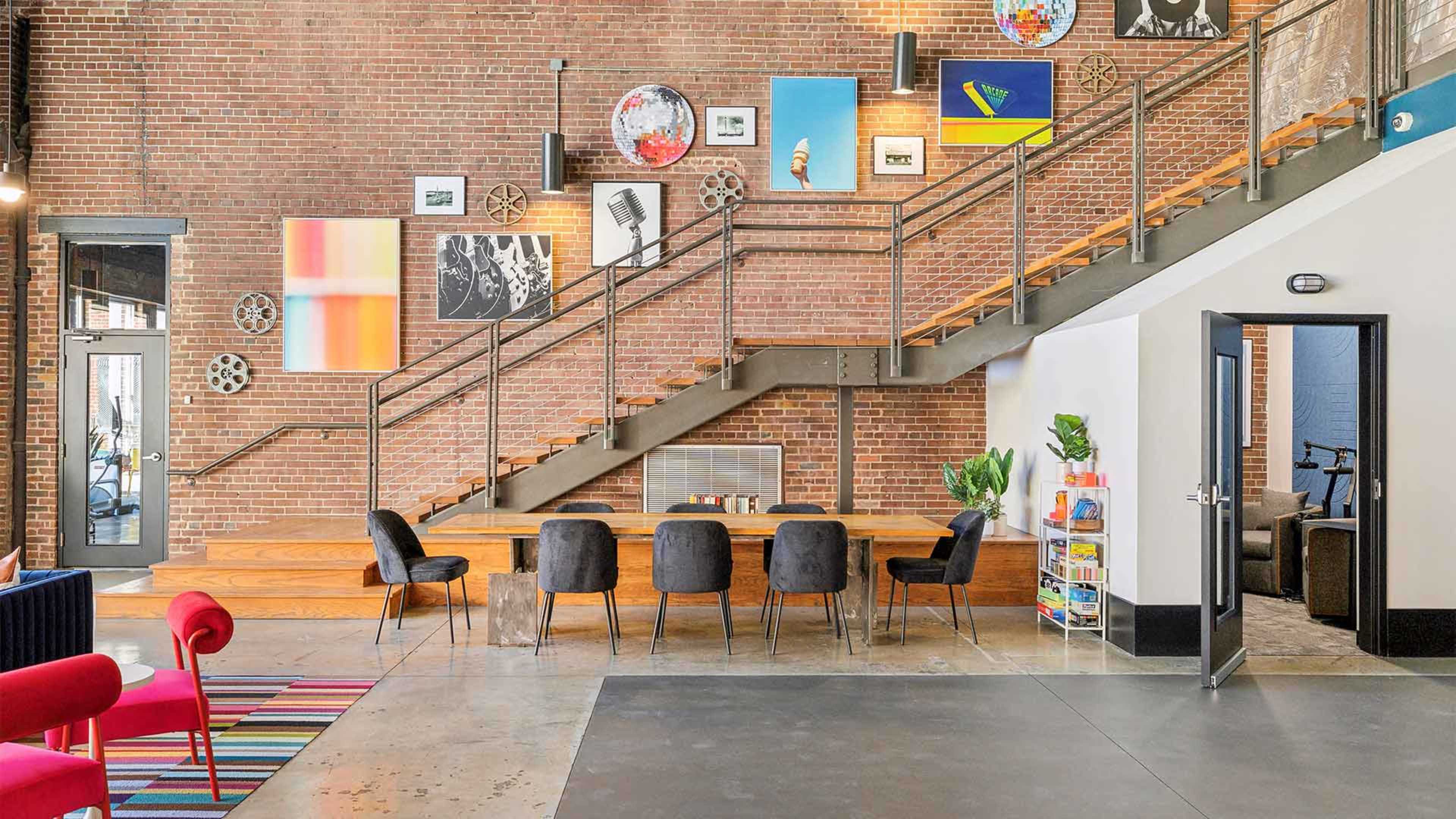 The image shows a modern interior space featuring a staircase, seating area with chairs, and walls adorned with various artworks.