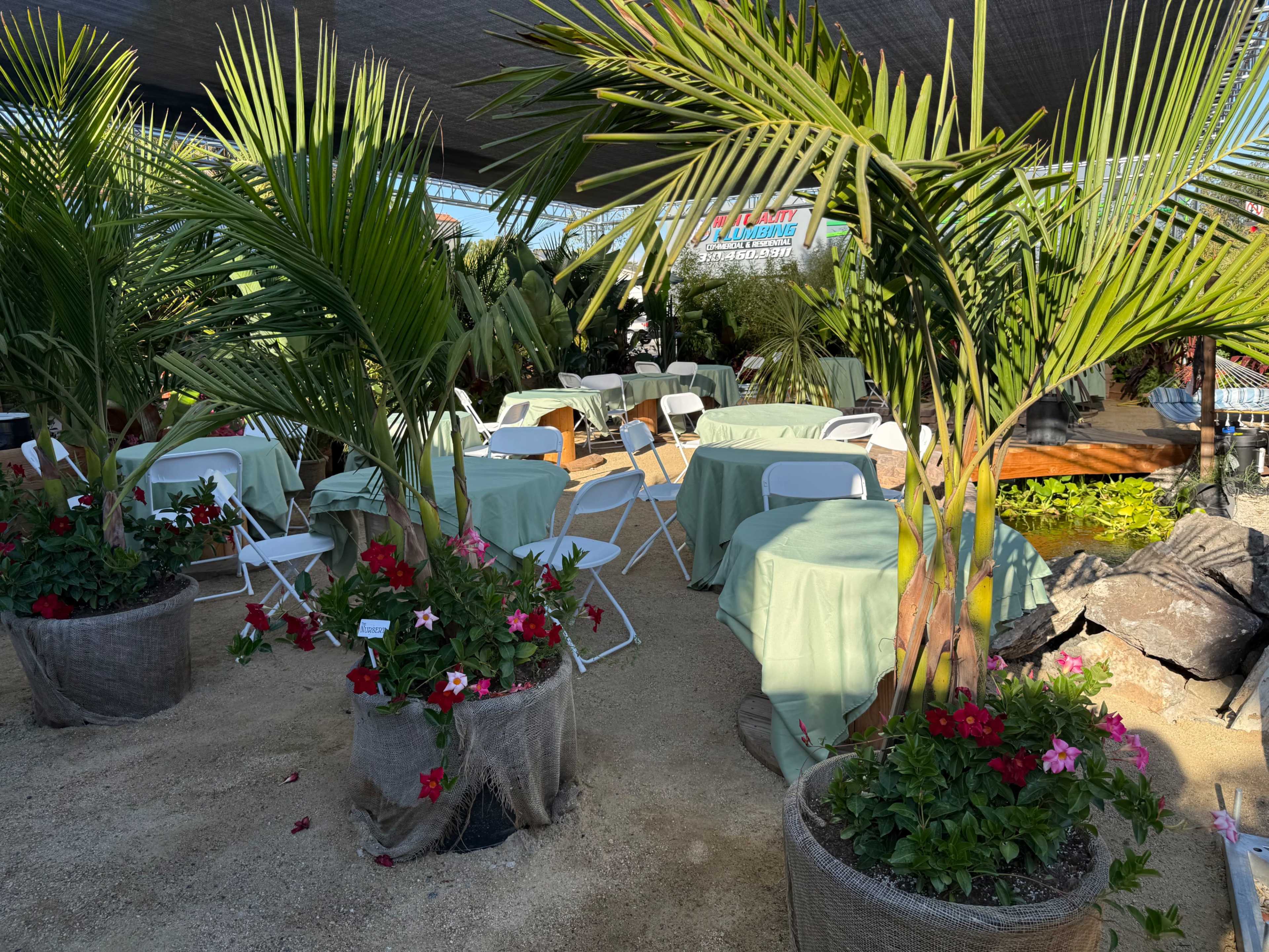 The scene features a garden area with white chairs and tables covered in light green cloth, surrounded by potted plants and flowers.