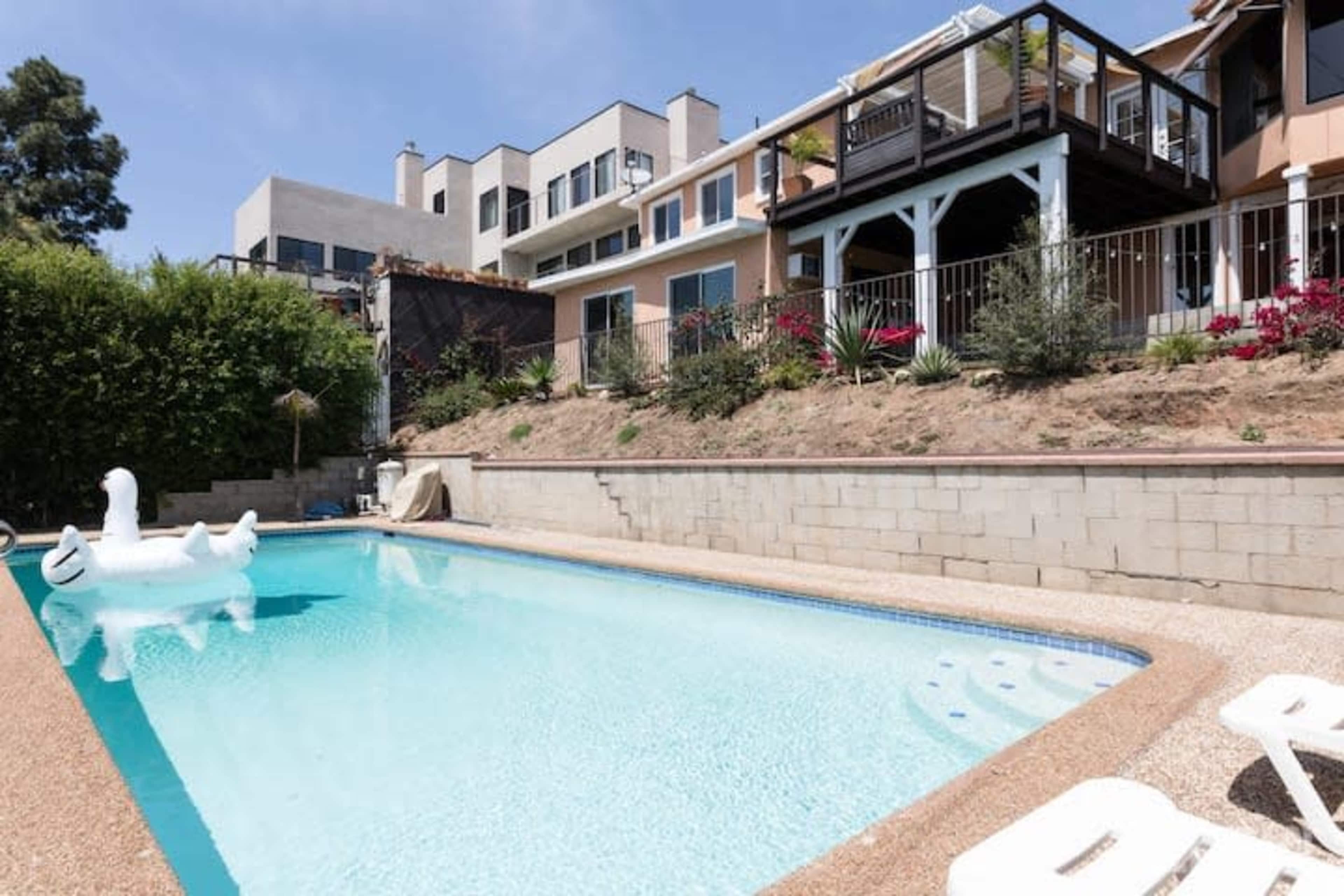 A swimming pool surrounded by a low stone wall, with residential buildings and landscaped areas in the background.
