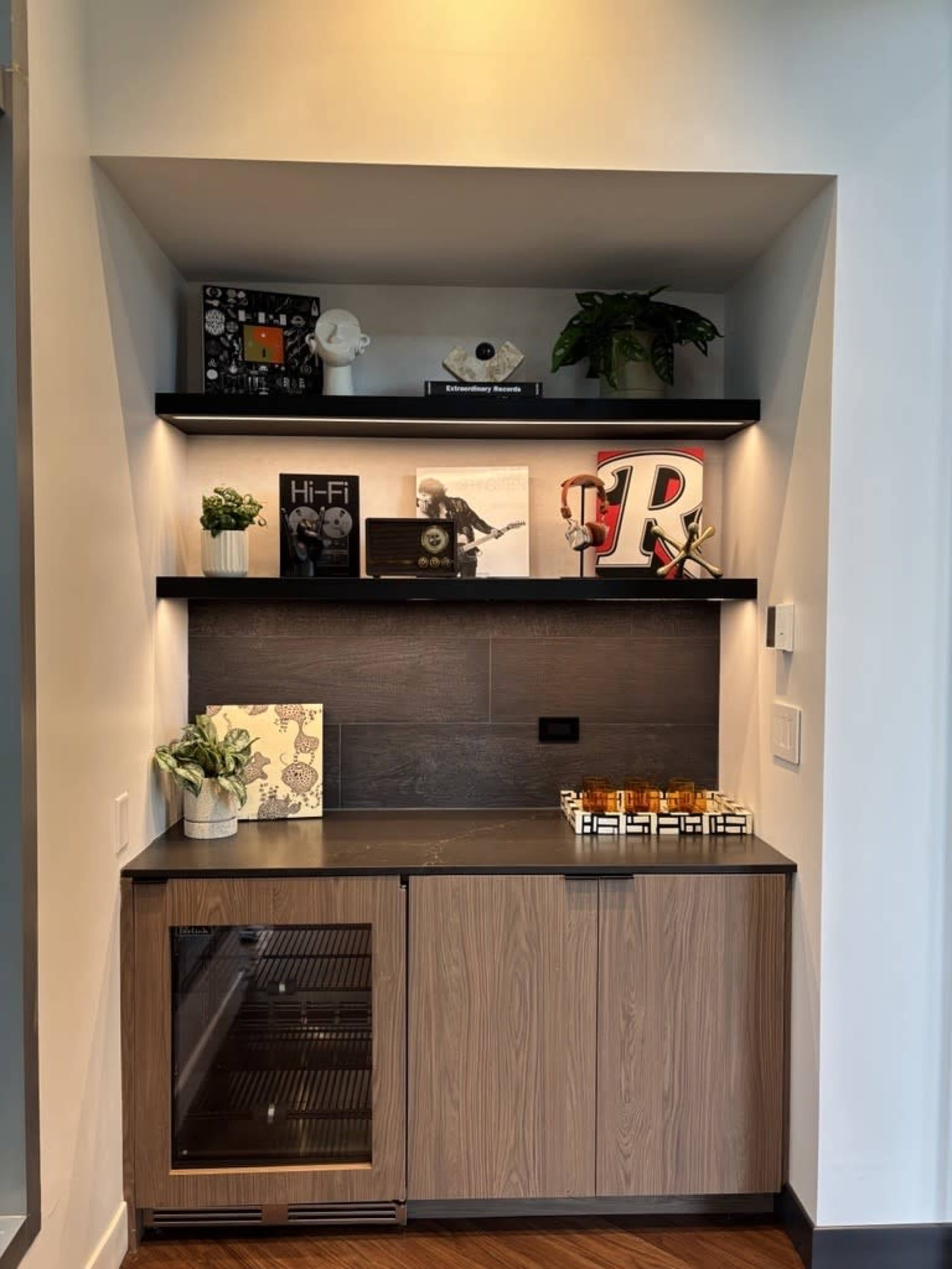 A modern nook features a dark countertop with an under-counter beverage cooler, surrounded by shelves displaying decorative items, plants, and framed photographs.