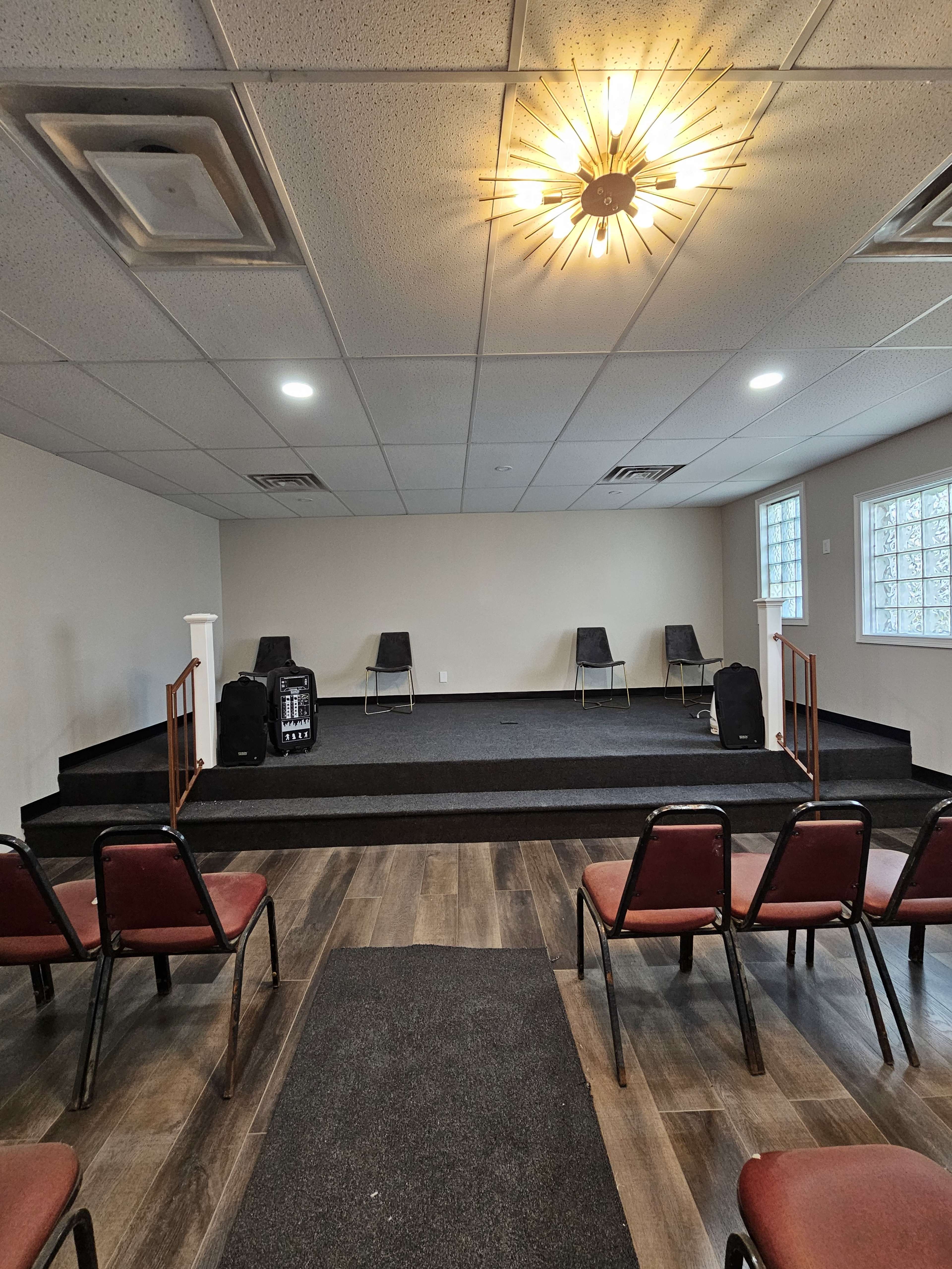 The image shows a small performance stage with four chairs arranged on it, flanked by a few bags, and facing a row of red upholstered chairs set up for an audience.