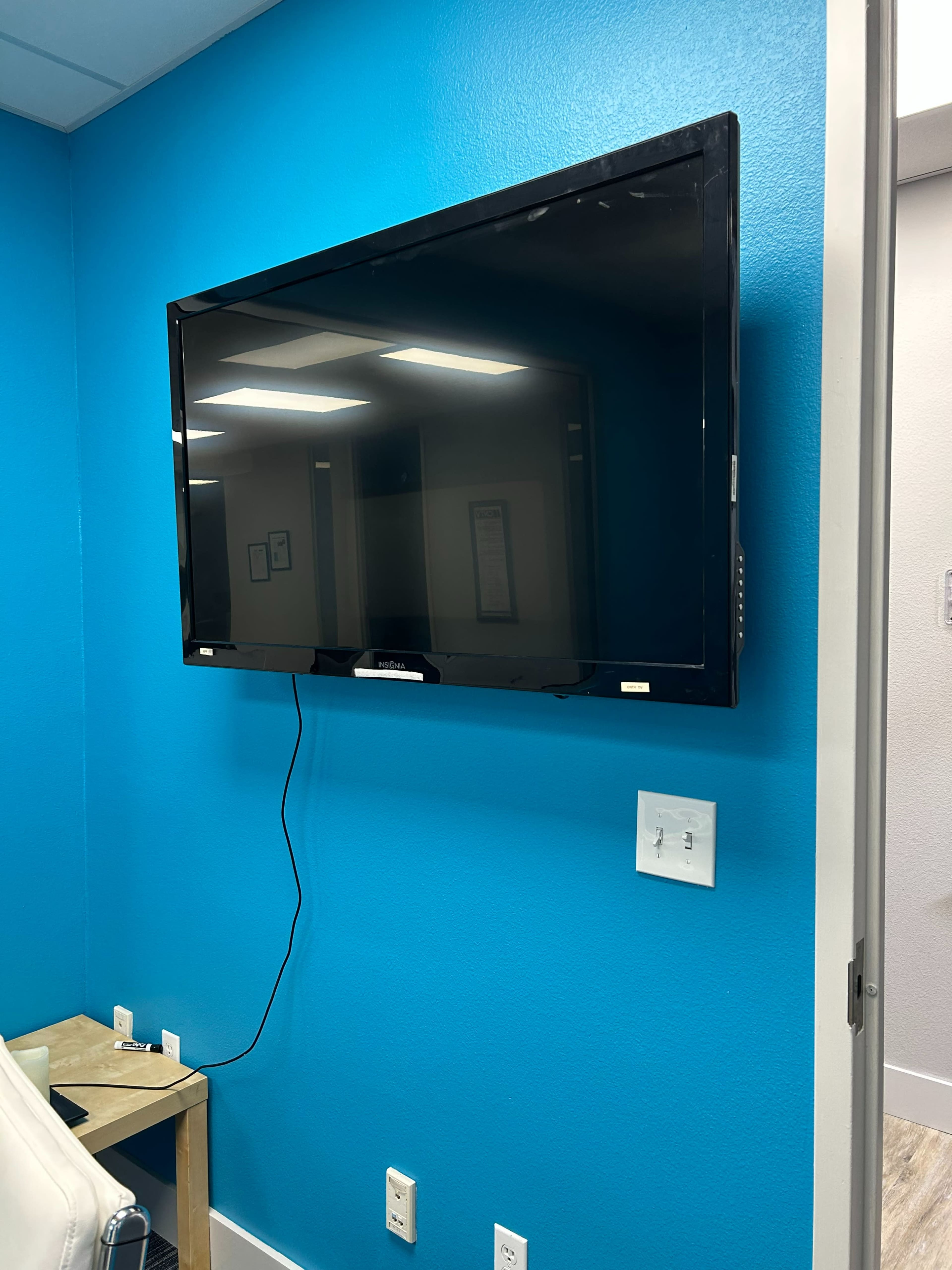 A flat-screen television is mounted on a bright blue wall in a room with a wooden table and visible electrical outlets.