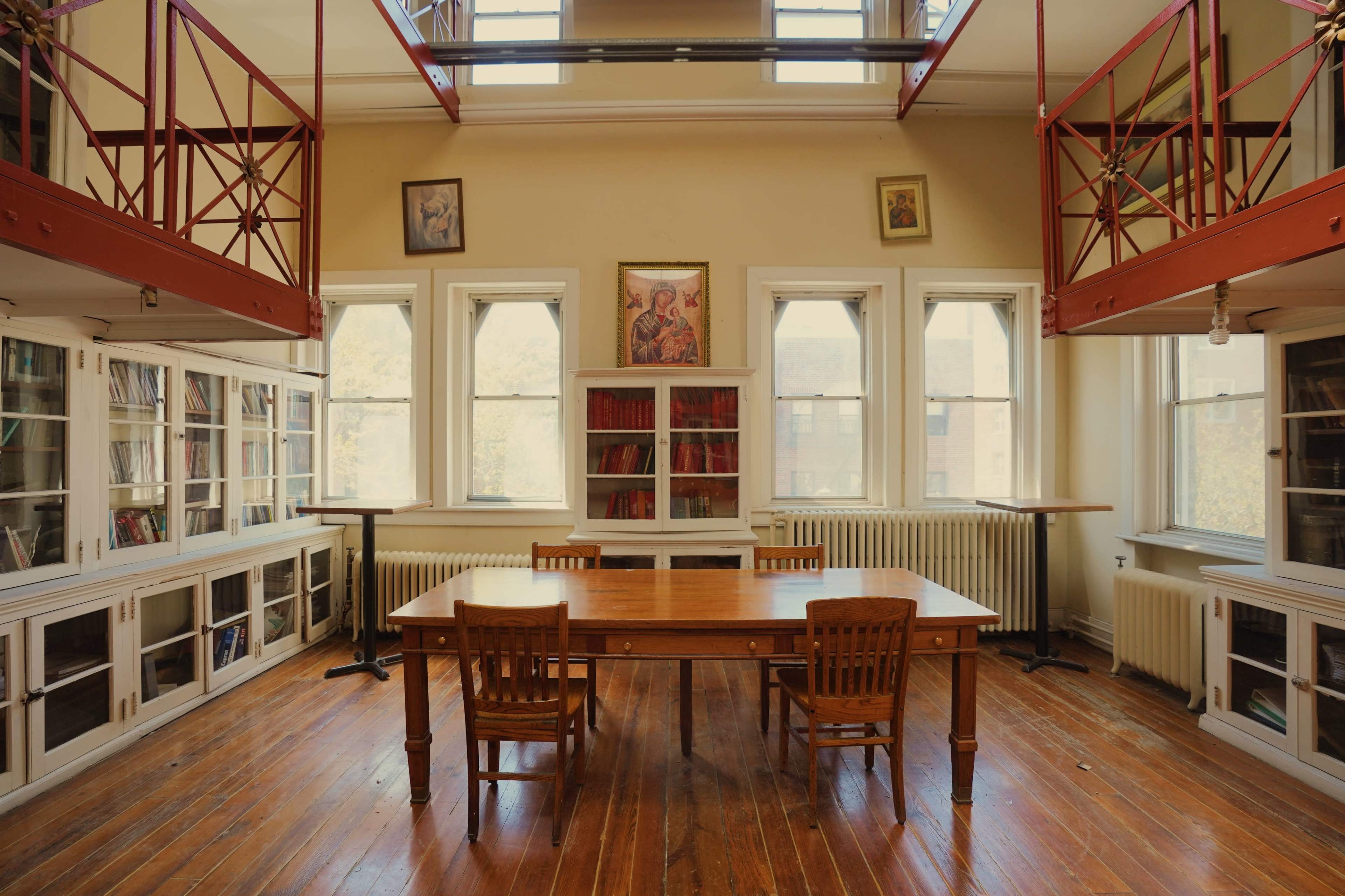 A spacious library room with a central wooden table and two chairs, surrounded by bookshelves and large windows.