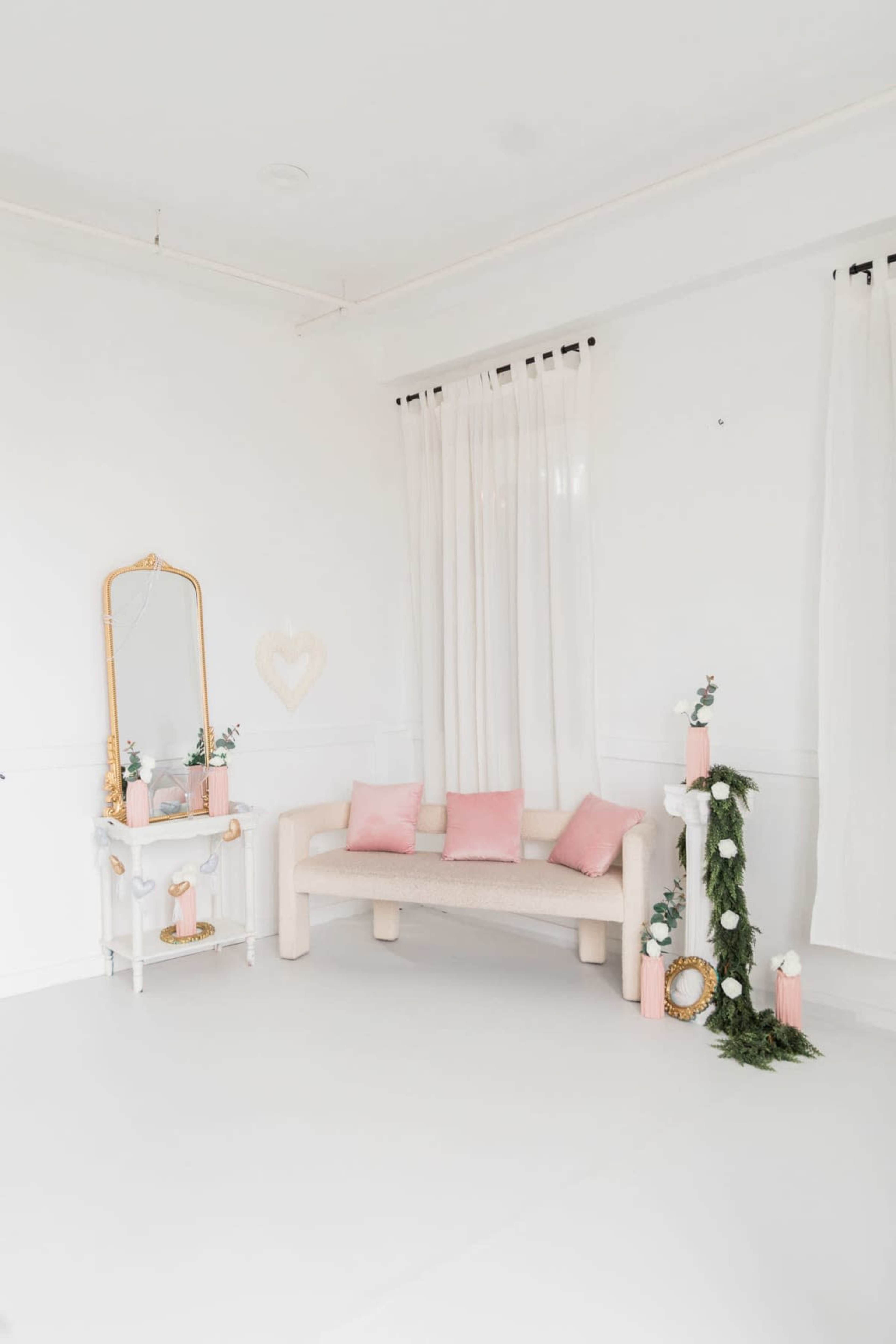 The image shows a minimalistic white room with a light-colored sofa adorned with pink cushions, a decorative mirror, and greenery with candles on a console table.