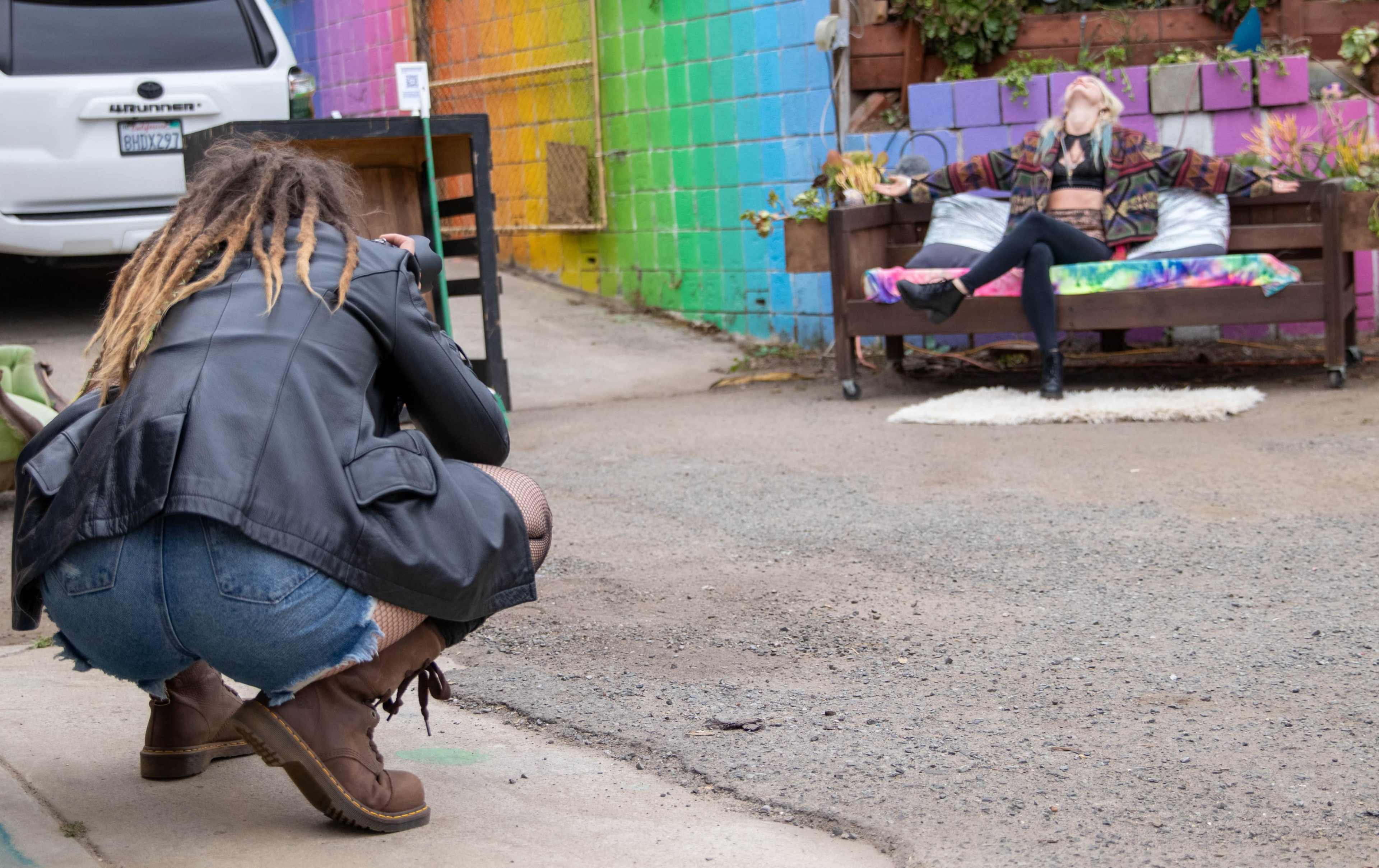 A photographer crouches on a street while capturing a model posing on a bench in front of a colorful mural.