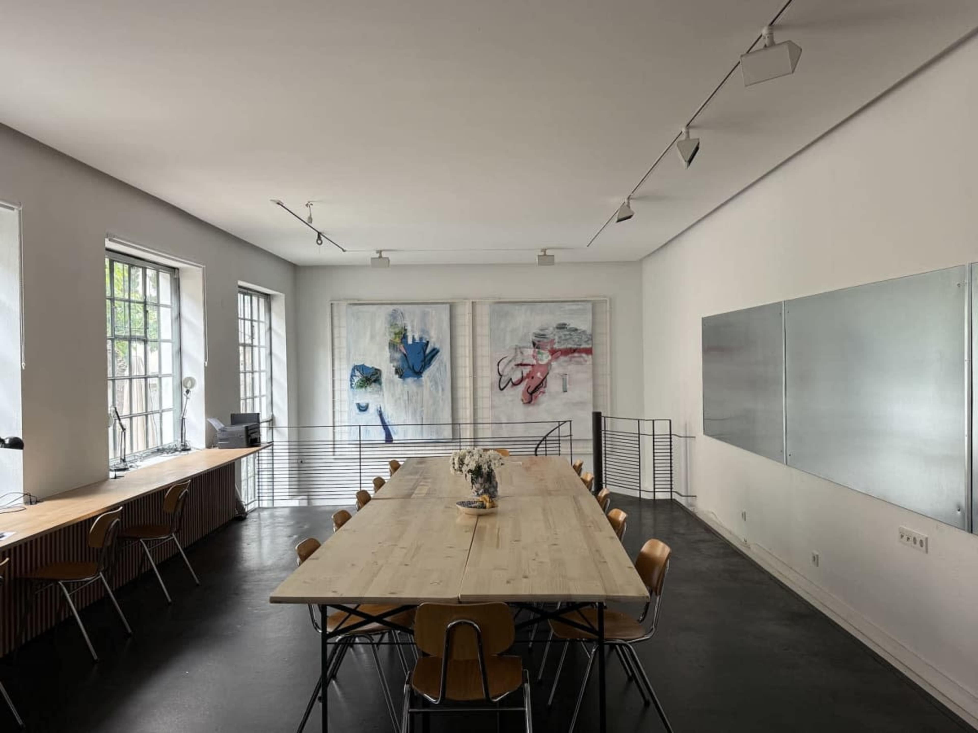 A spacious meeting room features a long wooden table surrounded by chairs, with large windows and abstract artwork on the walls.