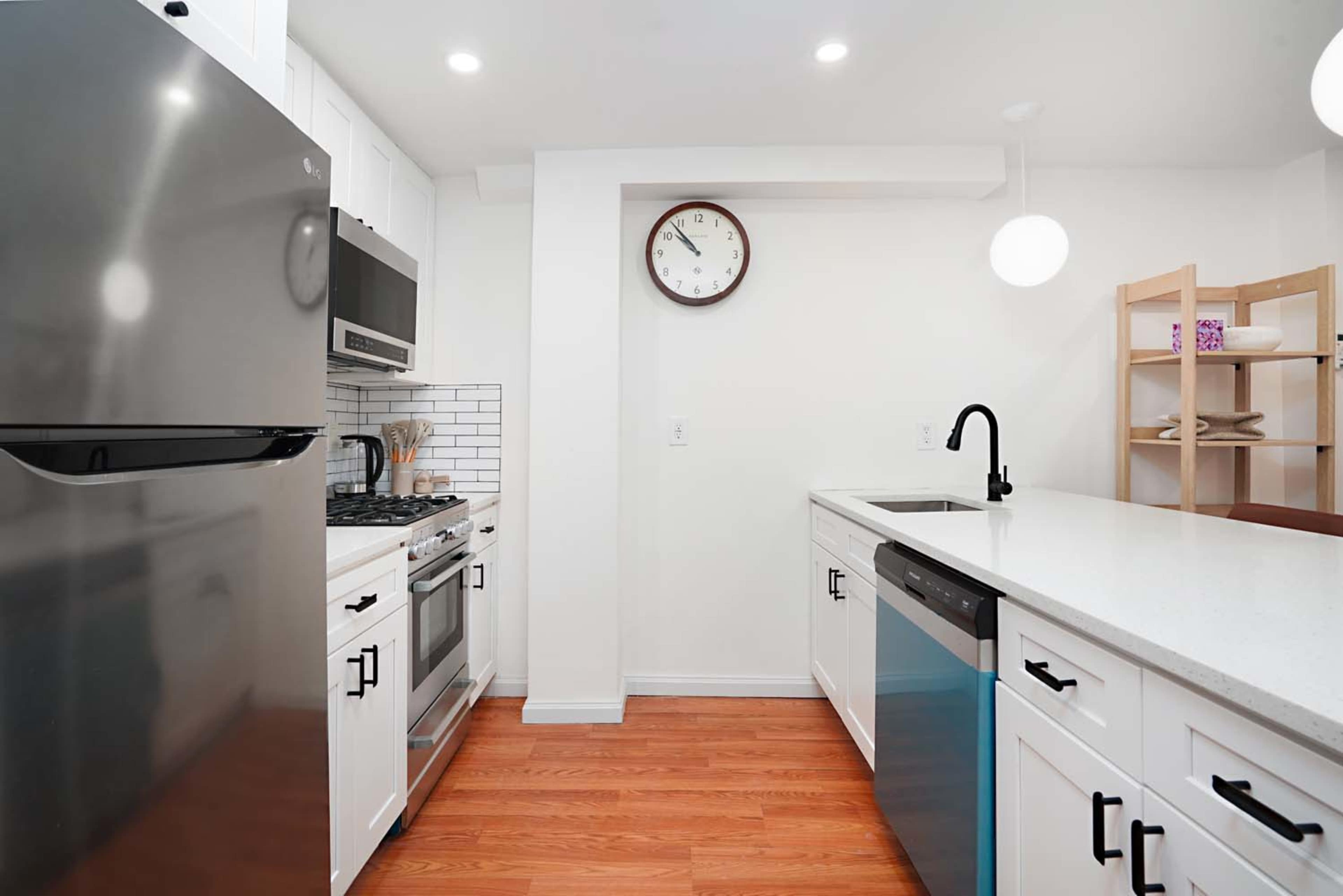 The image shows a modern kitchen featuring stainless steel appliances, white cabinetry, and a large quartz countertop with a sink and a wooden shelf.