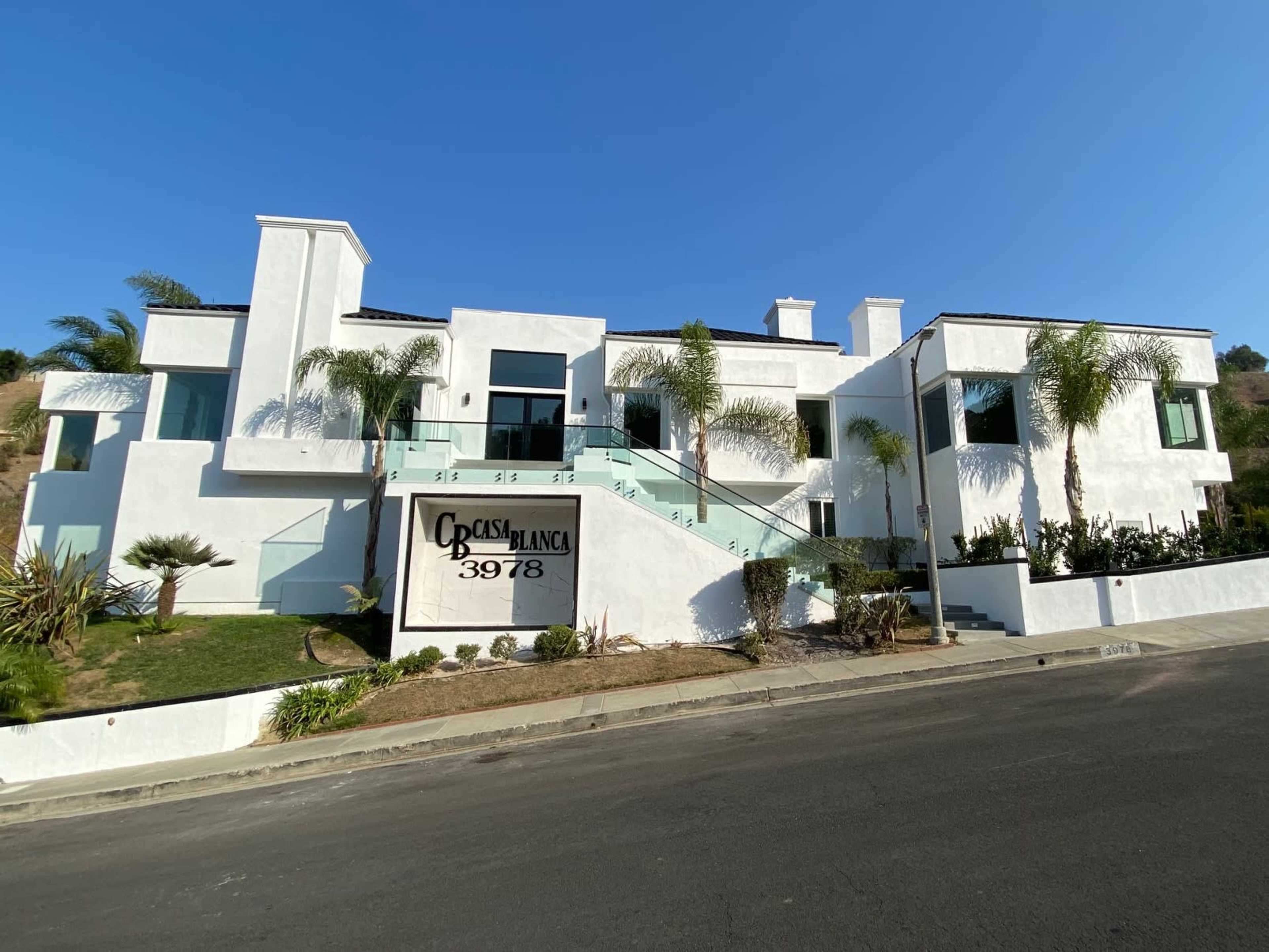 A modern white house with large windows and palm trees is situated on a sloped lot.