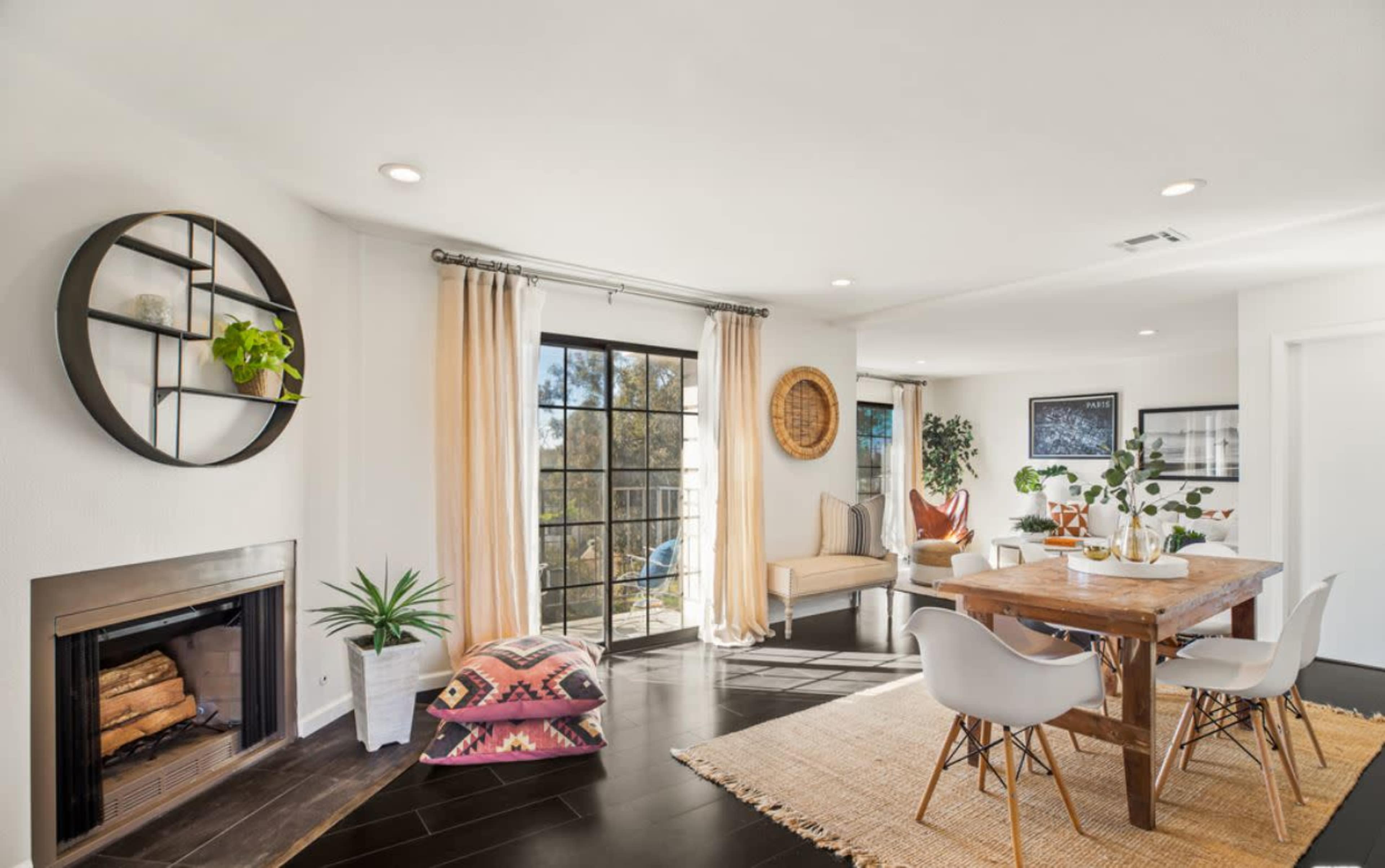 A light-filled living space featuring a dining area with a wooden table, white chairs, a fireplace, and large windows leading to an outdoor view.