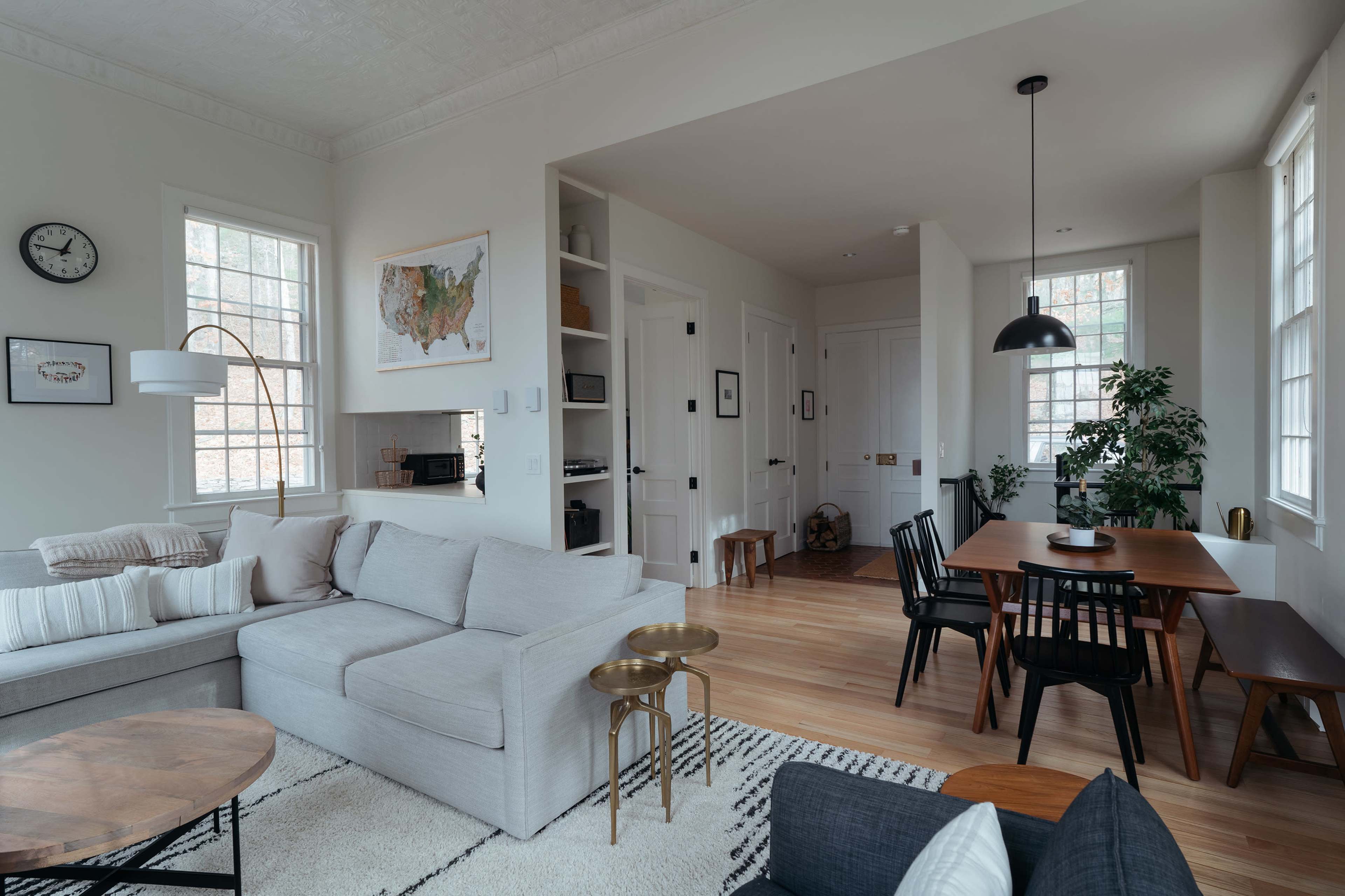 A modern living room with a light-colored sofa, a round coffee table, a dining area with a wooden table and chairs, and large windows allowing natural light to enter.