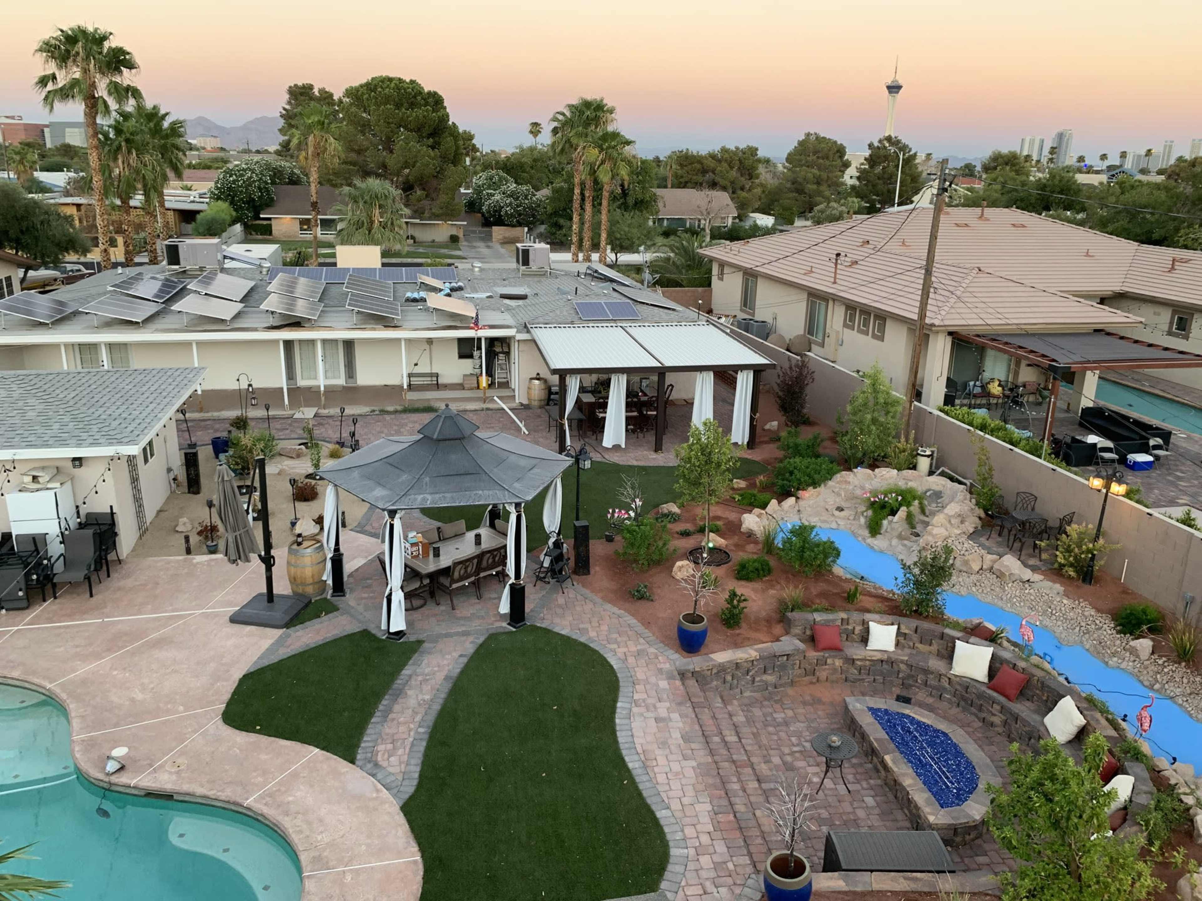 The image shows a backyard with a swimming pool, landscaped areas, a gazebo, a stone patio, and several palm trees under a sunset sky.
