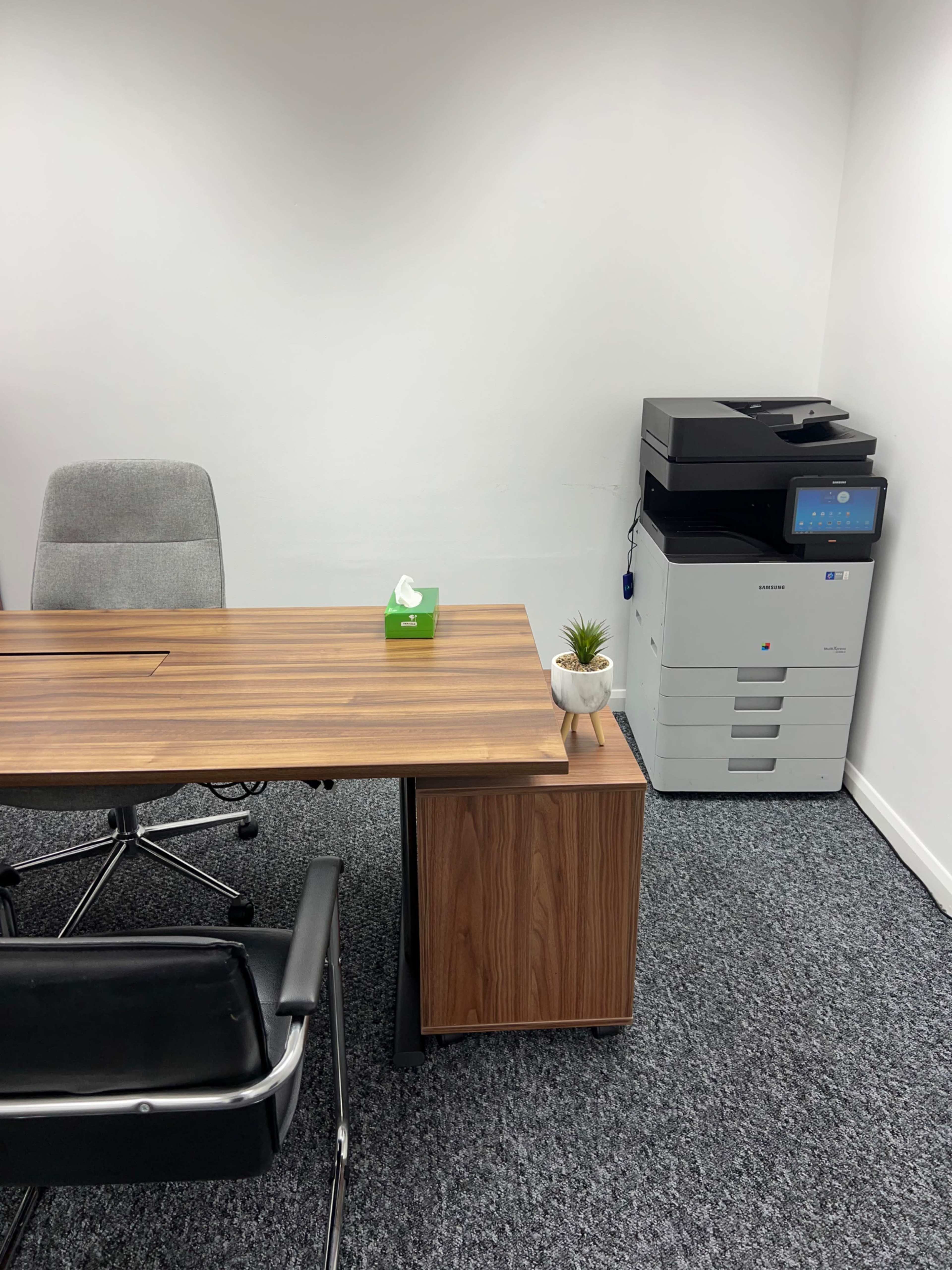 The image shows an office space with a wooden desk and rolling chair, a green tissue box, a small plant in a pot, and a photocopier in the corner.