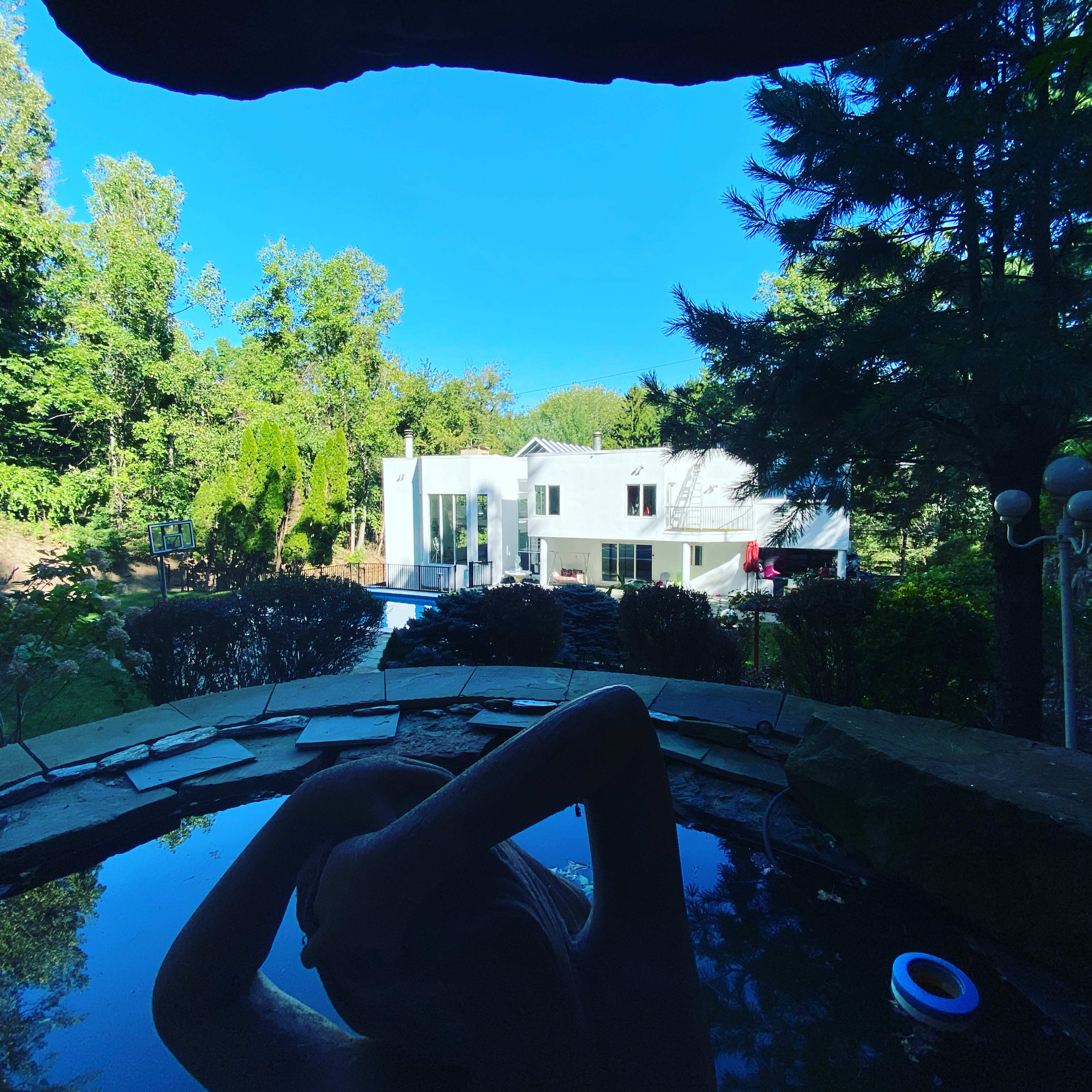 A person relaxes in a hot tub while overlooking a modern house and lush greenery.