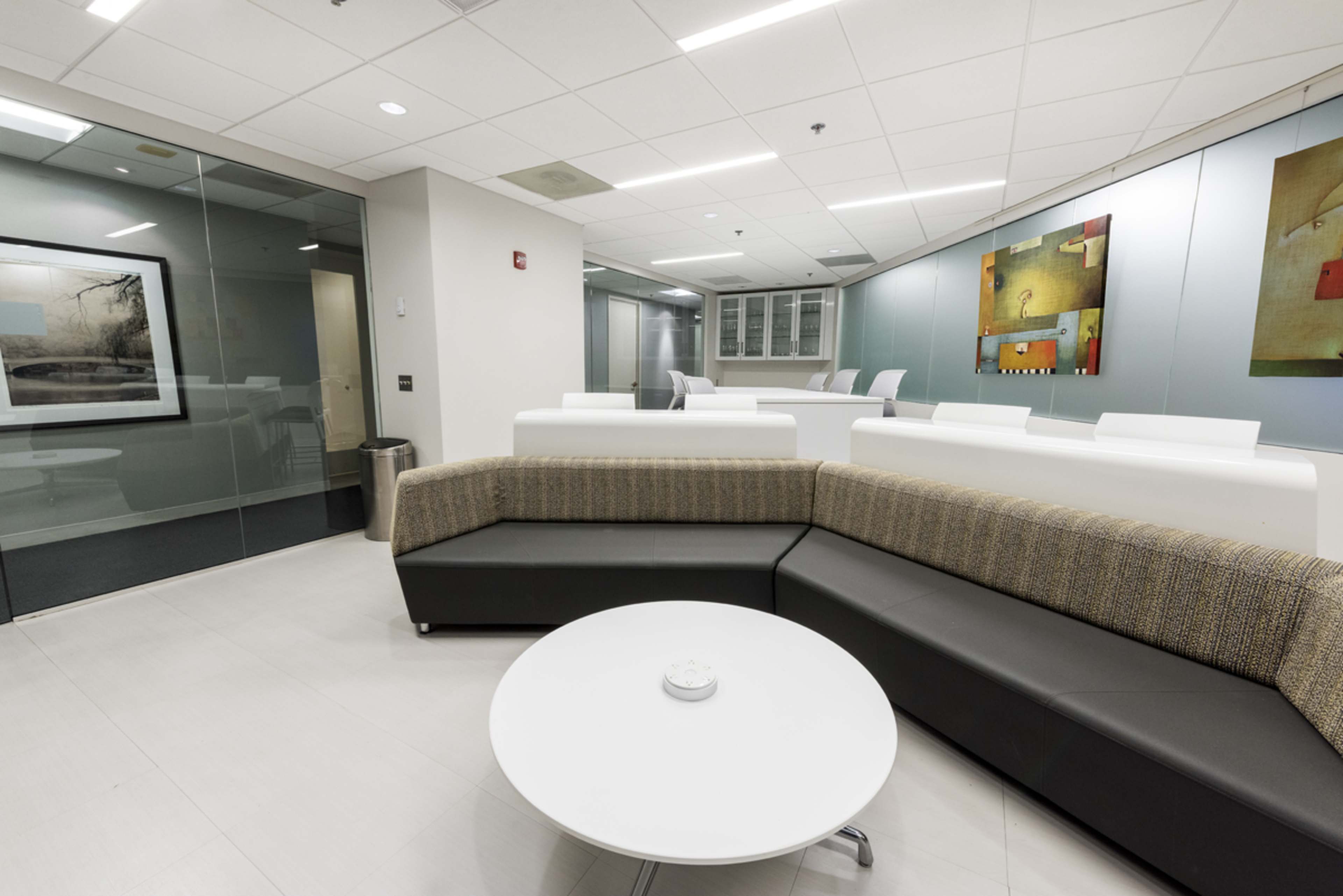 The image shows a modern office lounge area featuring a curved sectional sofa and a round coffee table, with large windows and abstract artwork on the walls.