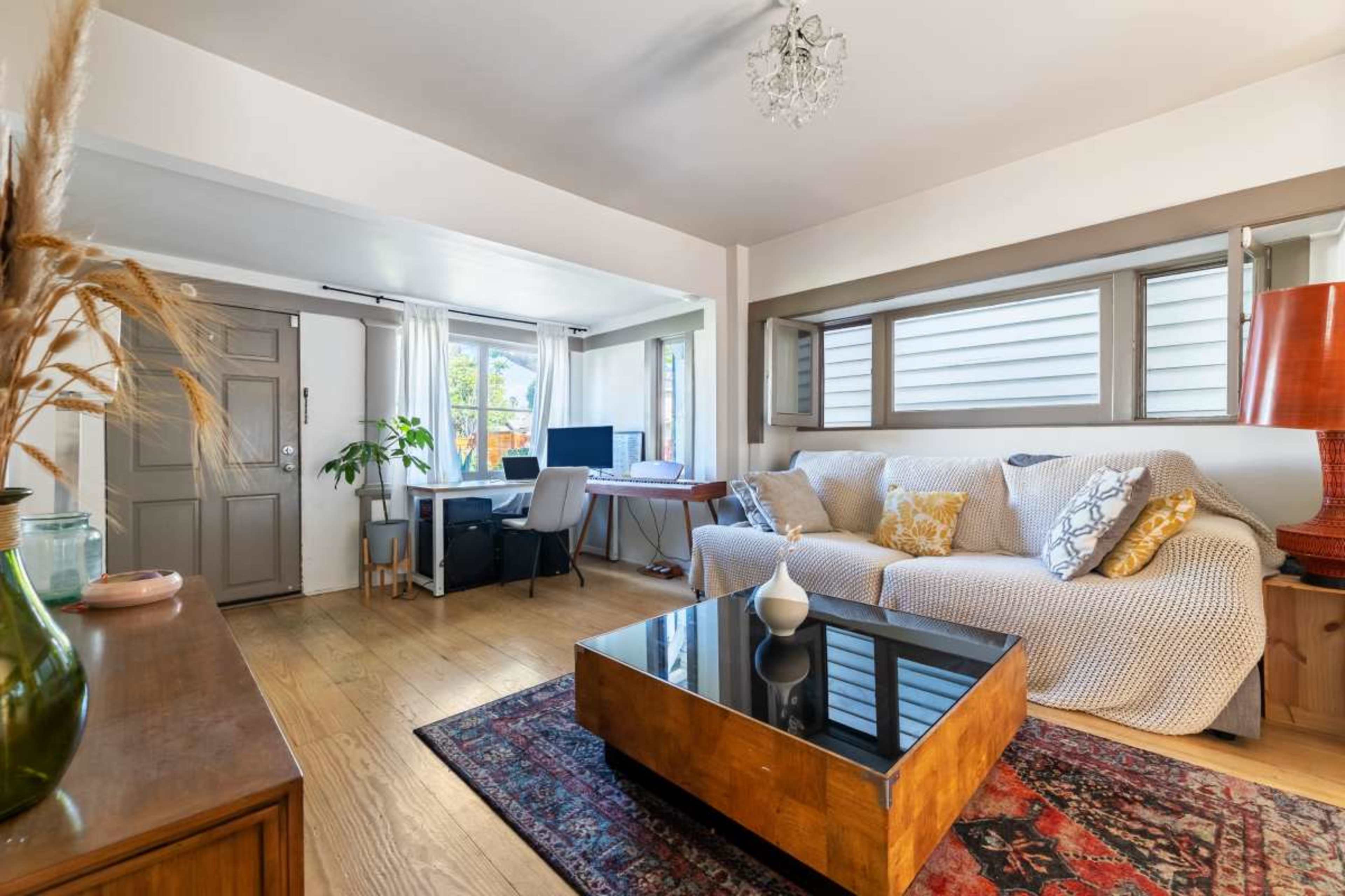 The image shows a living room with a sofa, a coffee table, a desk area, and large windows allowing natural light to enter.
