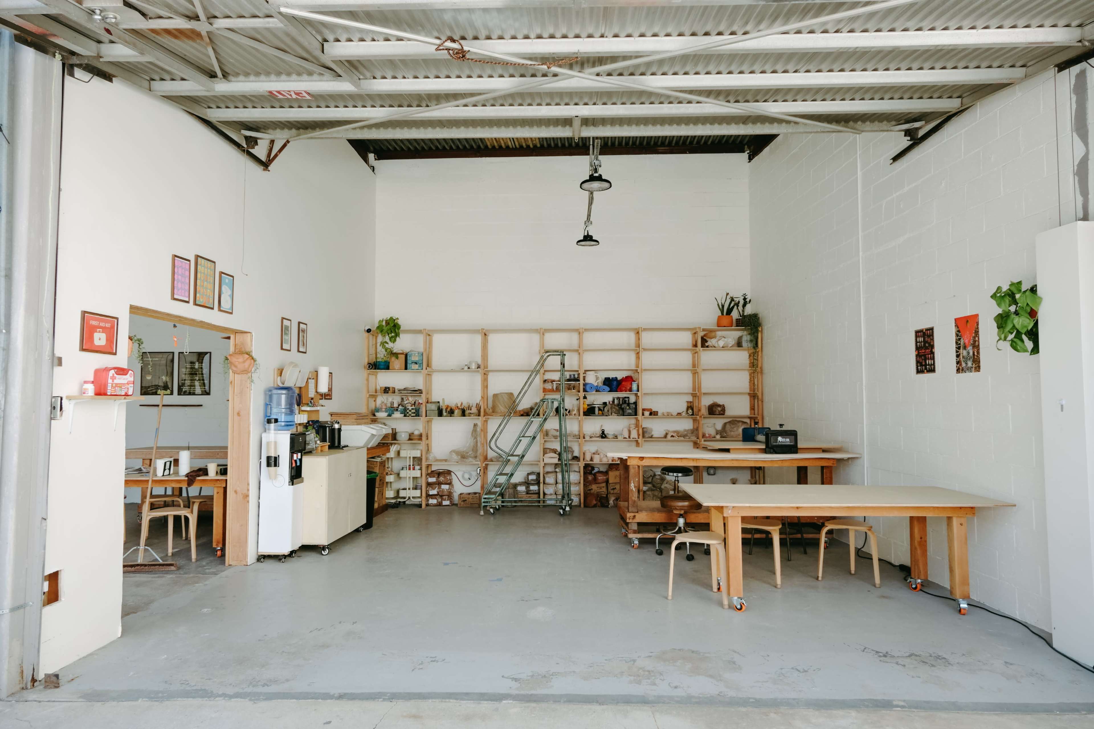 The image shows an empty, well-lit workspace featuring wooden shelves filled with items, a large table, chairs, and various appliances against a white wall.