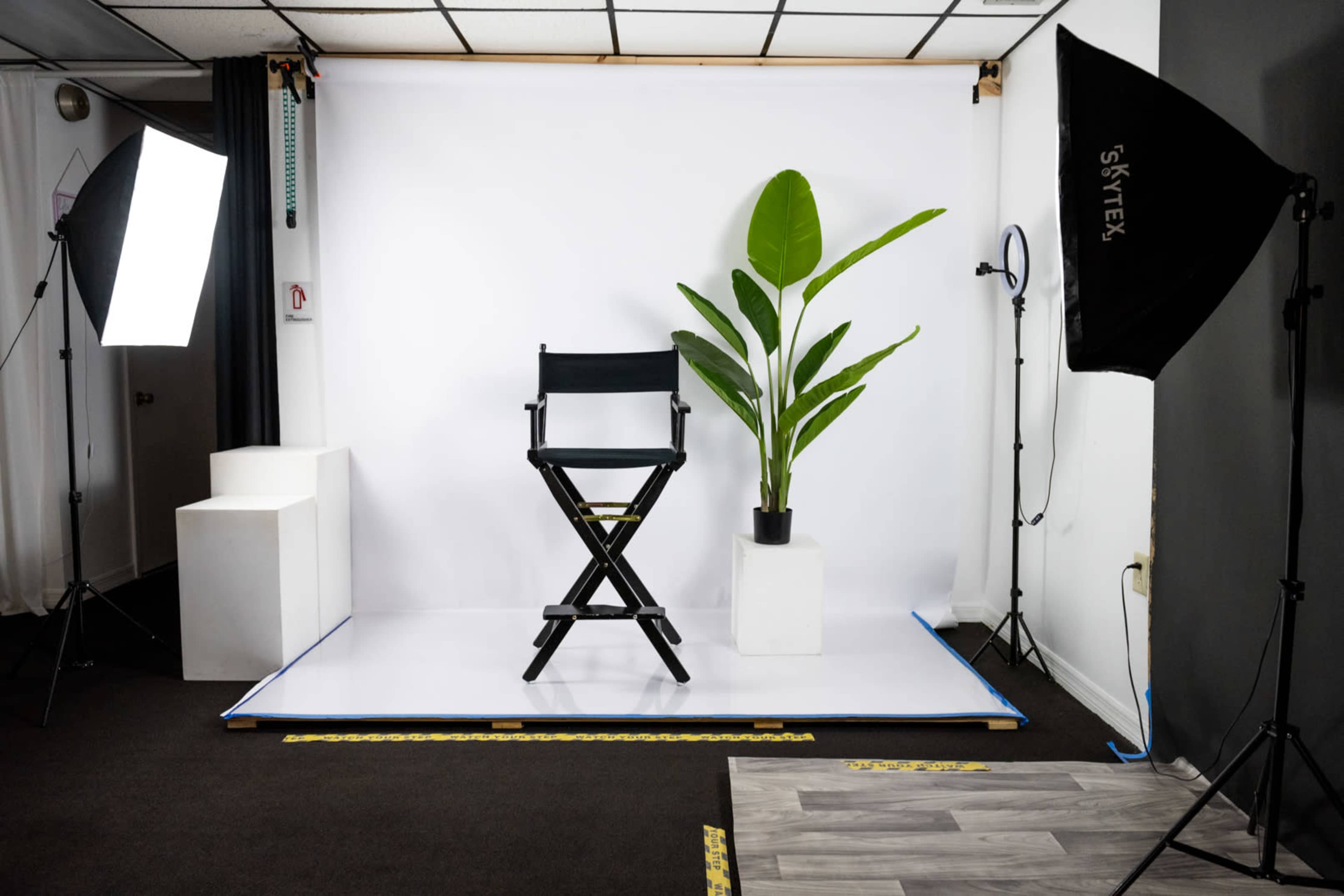 The image depicts a photography studio setup featuring a director's chair, a potted plant, and two softbox lights against a white backdrop.