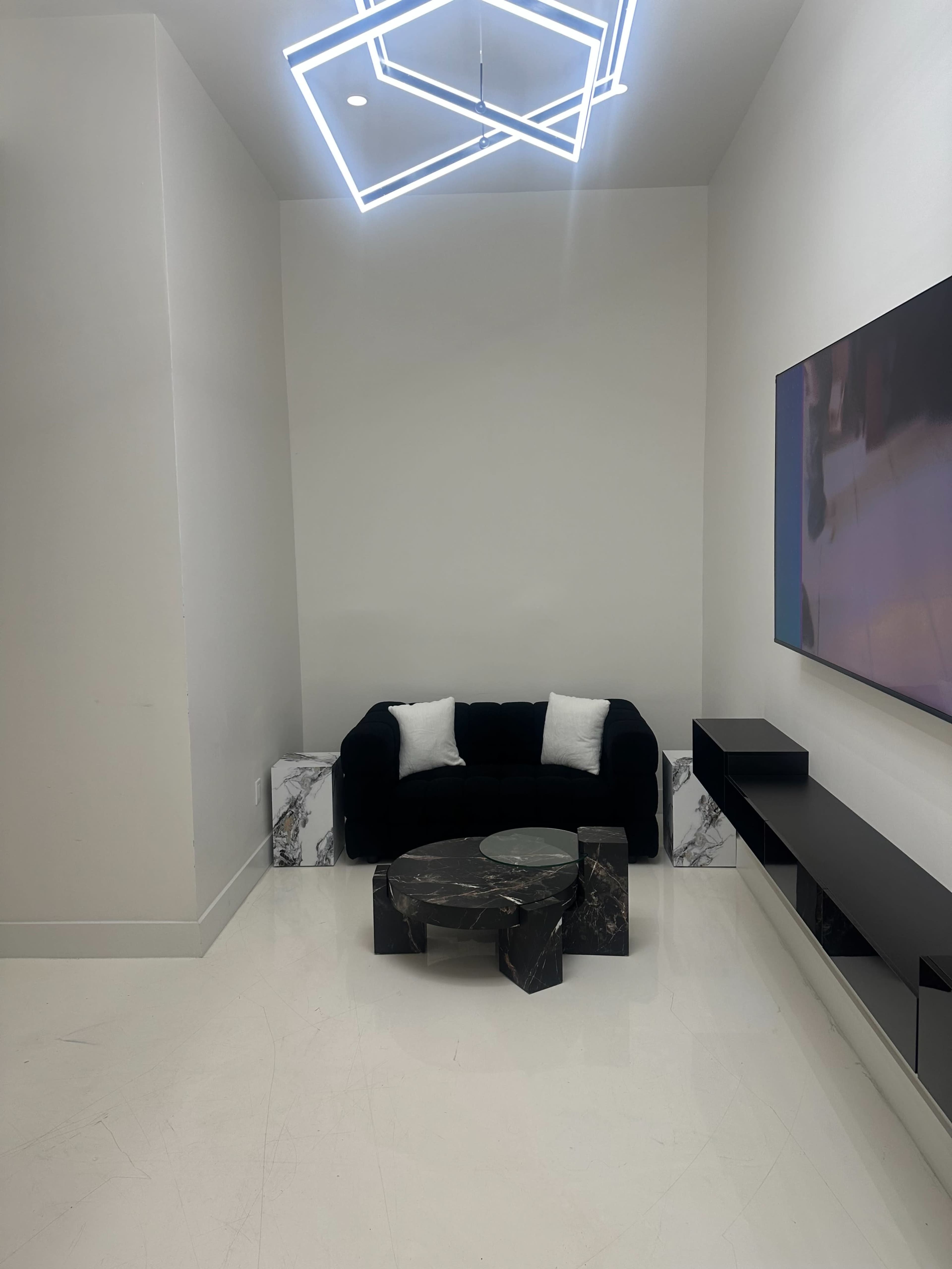 The image shows a modern living space with a dark velvet sofa, a circular coffee table, and a wall-mounted television under a geometric light fixture.