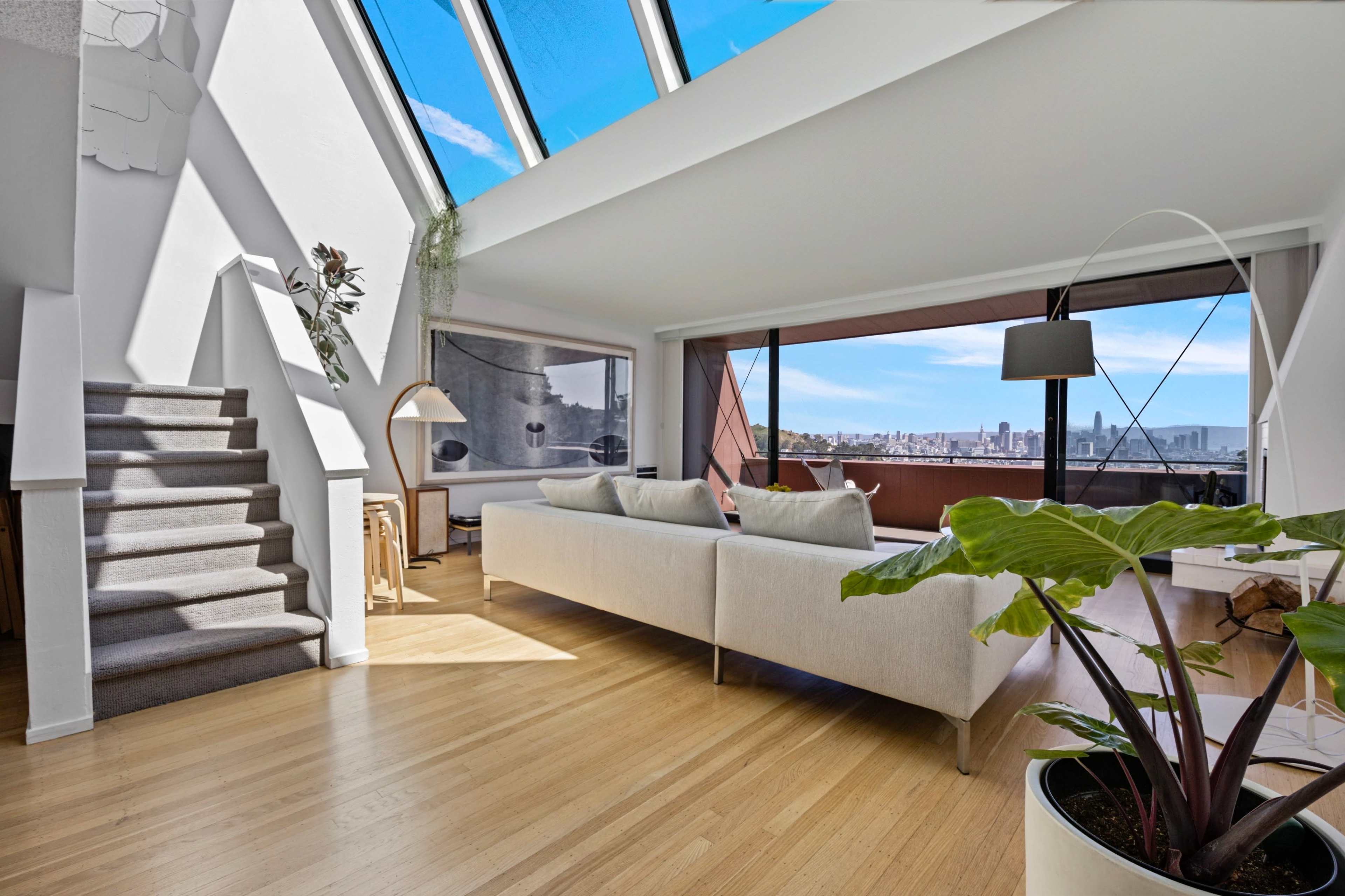The image shows a modern living room with a large skylight, a staircase, and a view of the city skyline through a wide glass window.