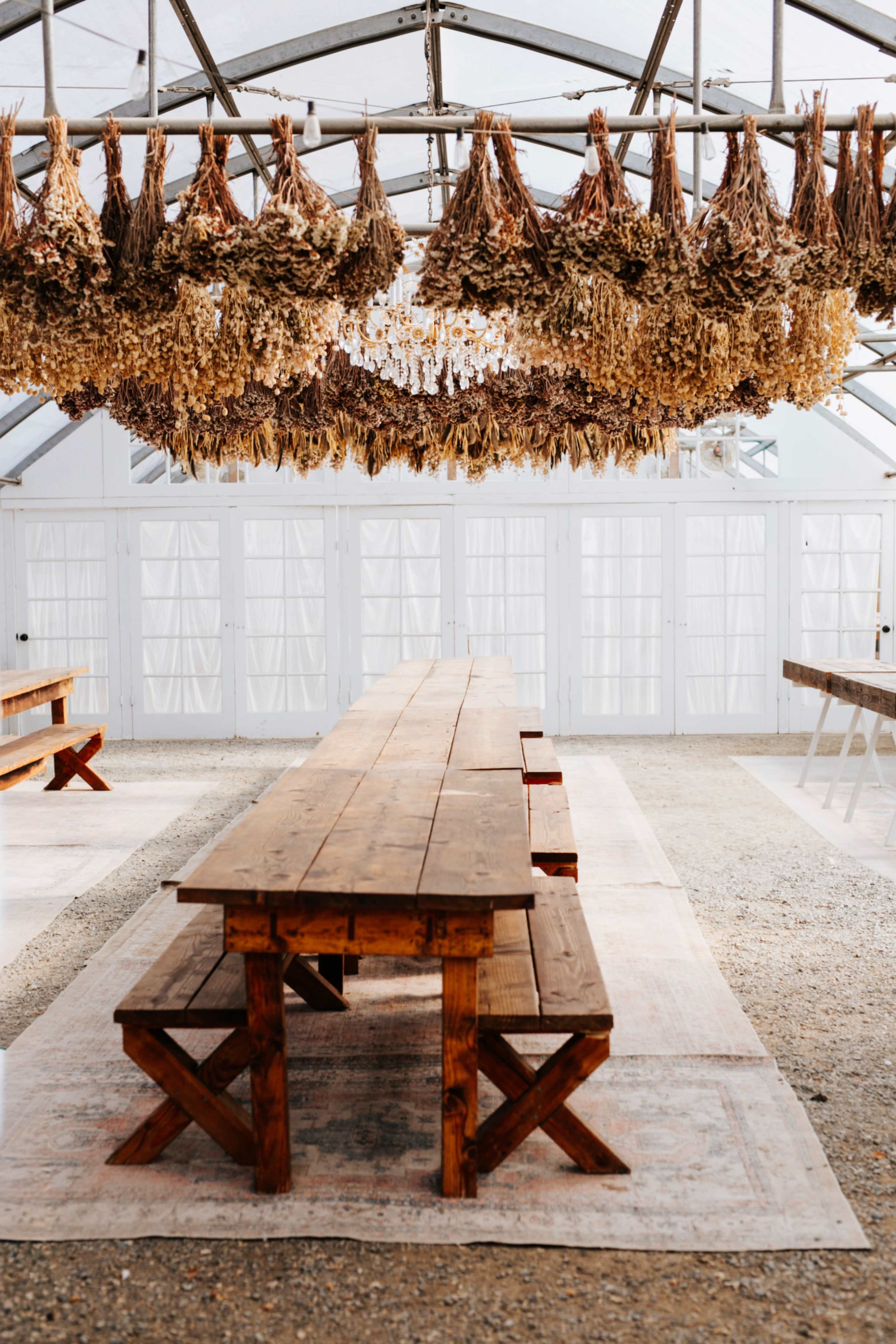The image shows a spacious indoor setting with long wooden tables beneath a structure adorned with hanging dried flowers.