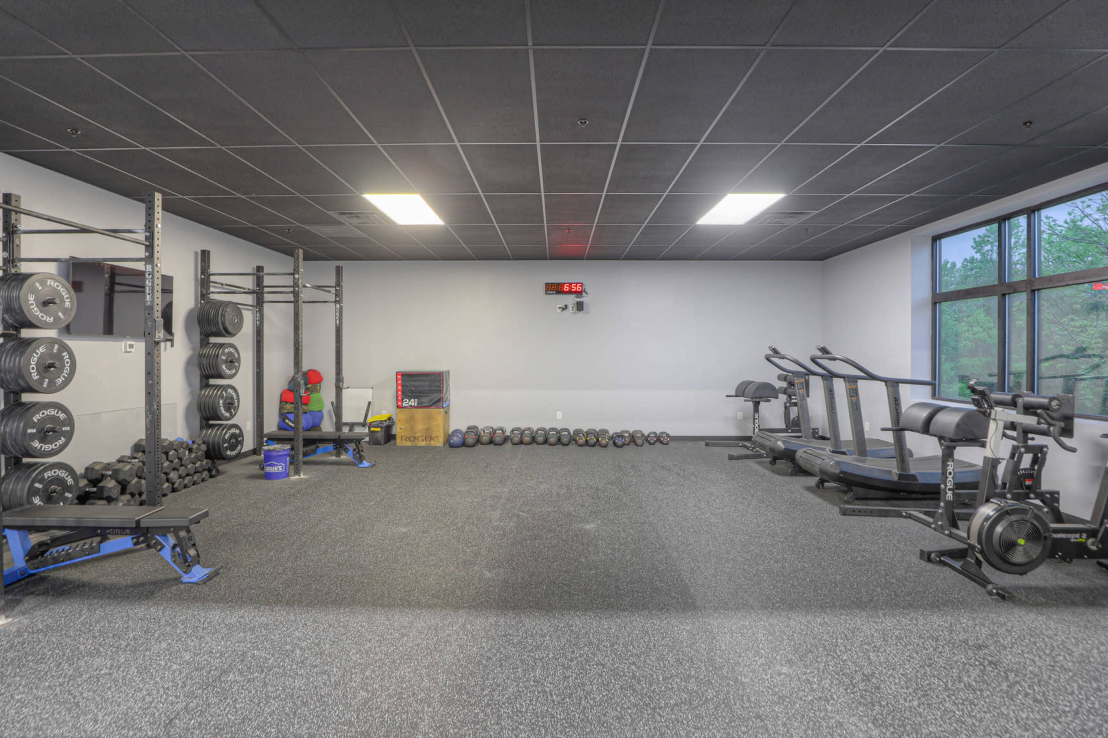 A fitness gym features weight racks, cardio machines, and a wall-mounted clock in a spacious, well-lit area.