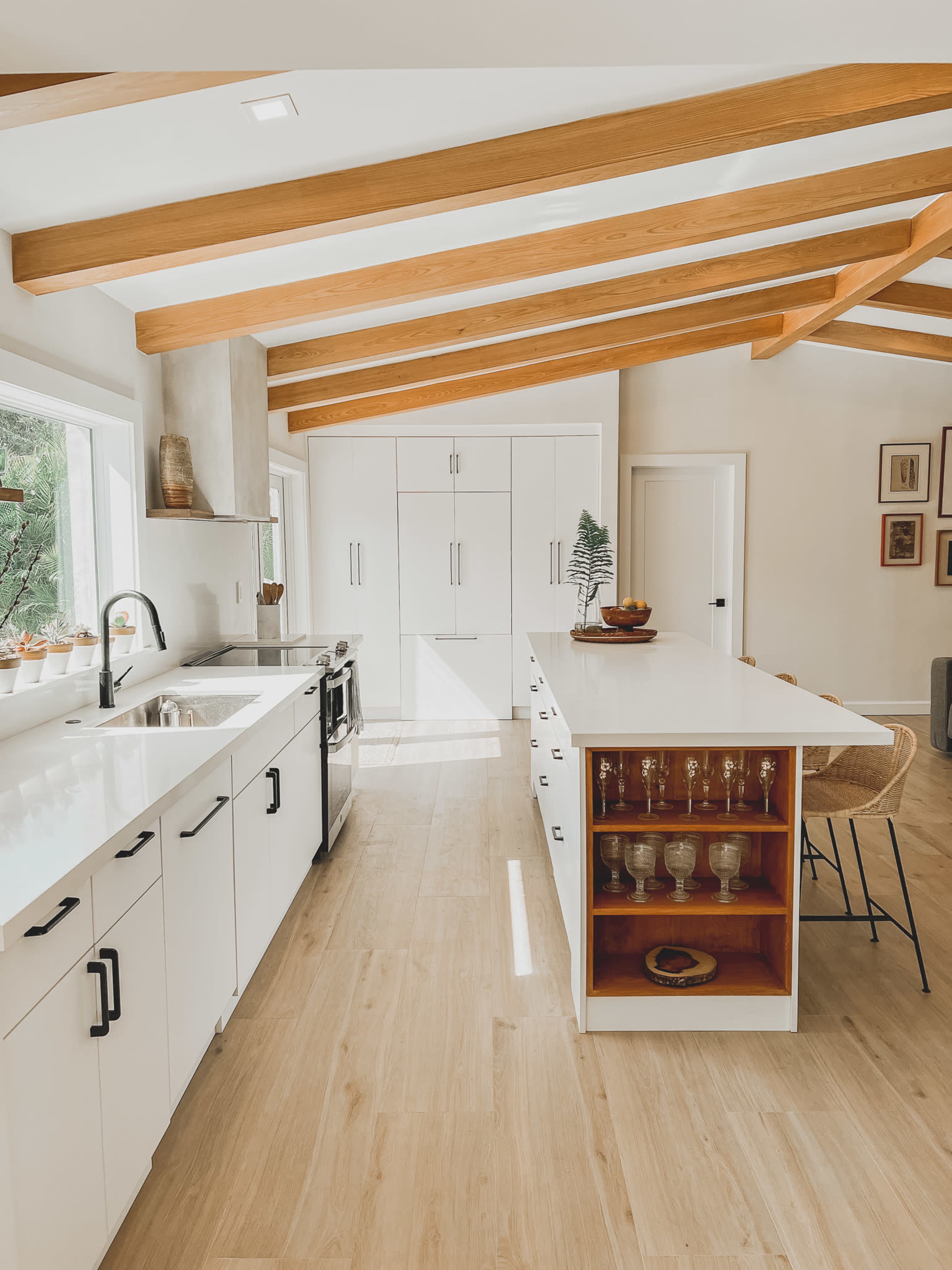 A bright kitchen features white cabinetry, wooden beams, and an island with open shelving displaying glassware.
