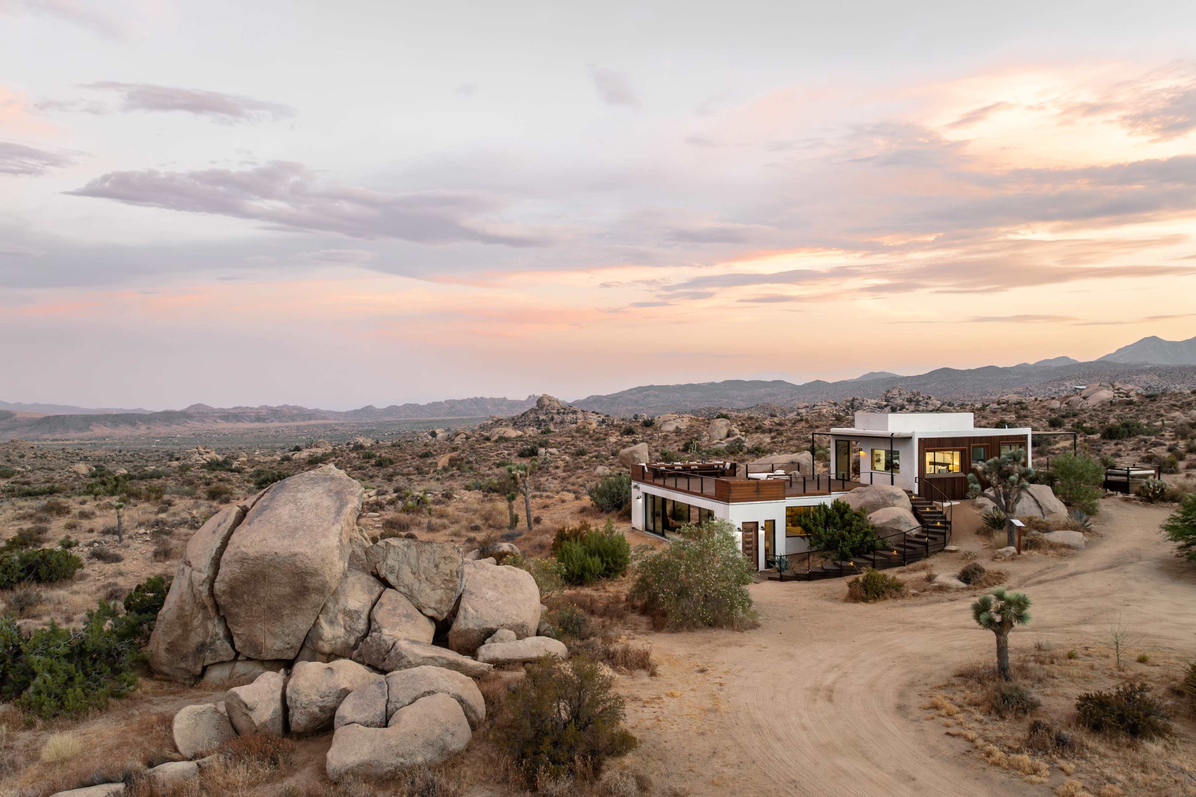 A modern house is situated among large boulders in a desert landscape under a colorful sunset sky.