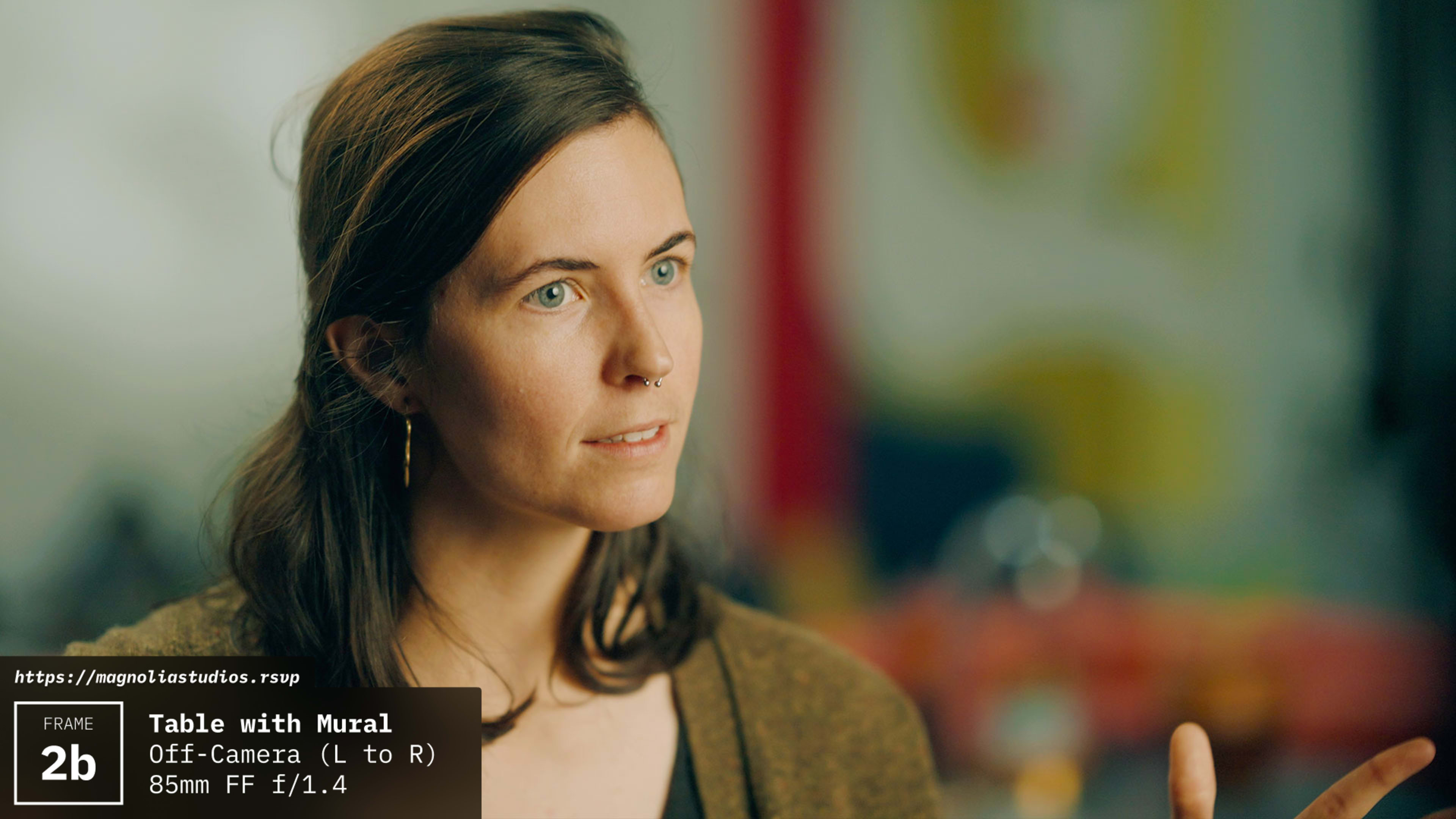 A woman gestures while speaking, with a colorful mural visible in the background.
