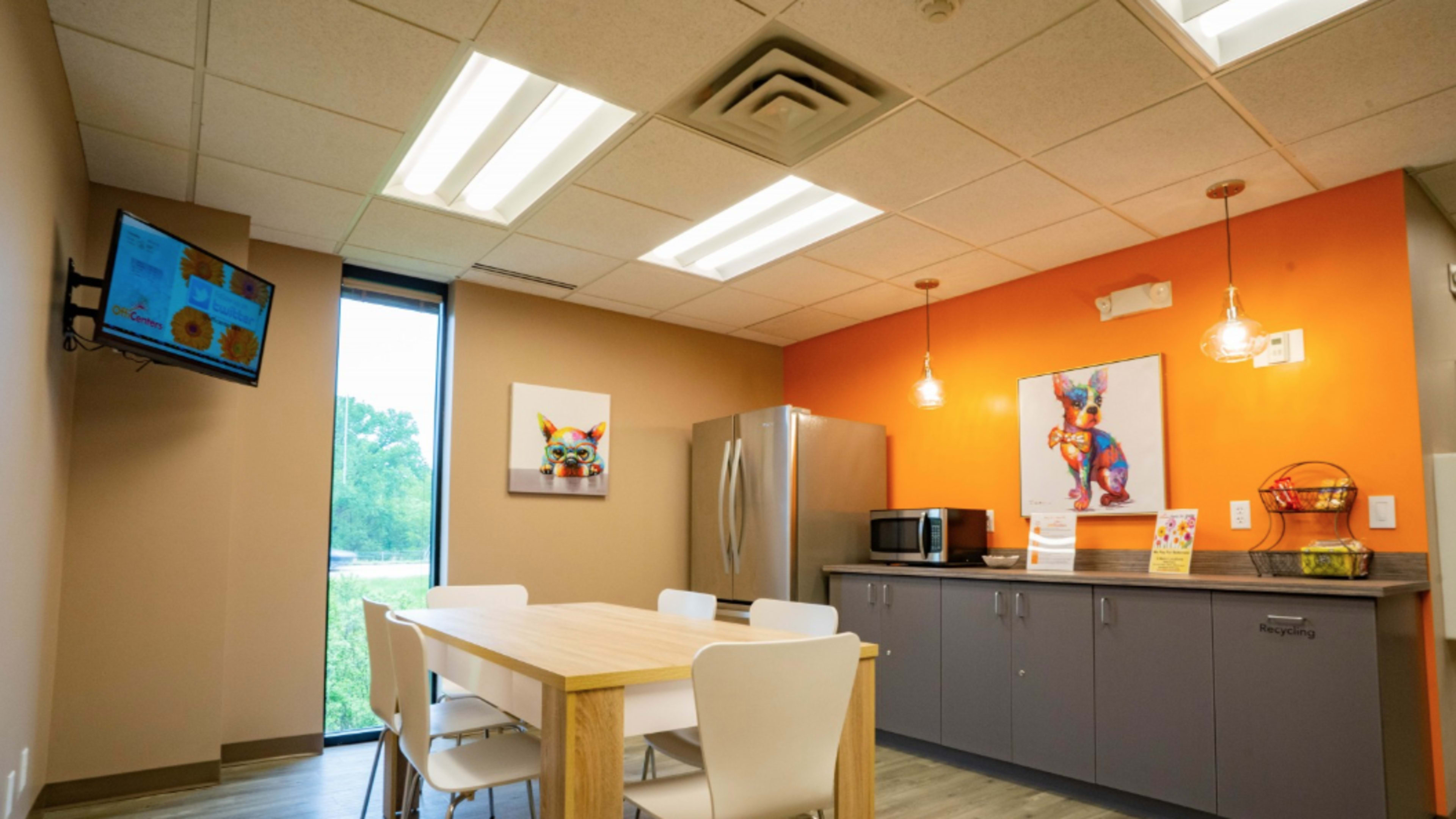 The image shows a modern break room with a wooden table, white chairs, a refrigerator, a microwave, and colorful artwork on the walls.