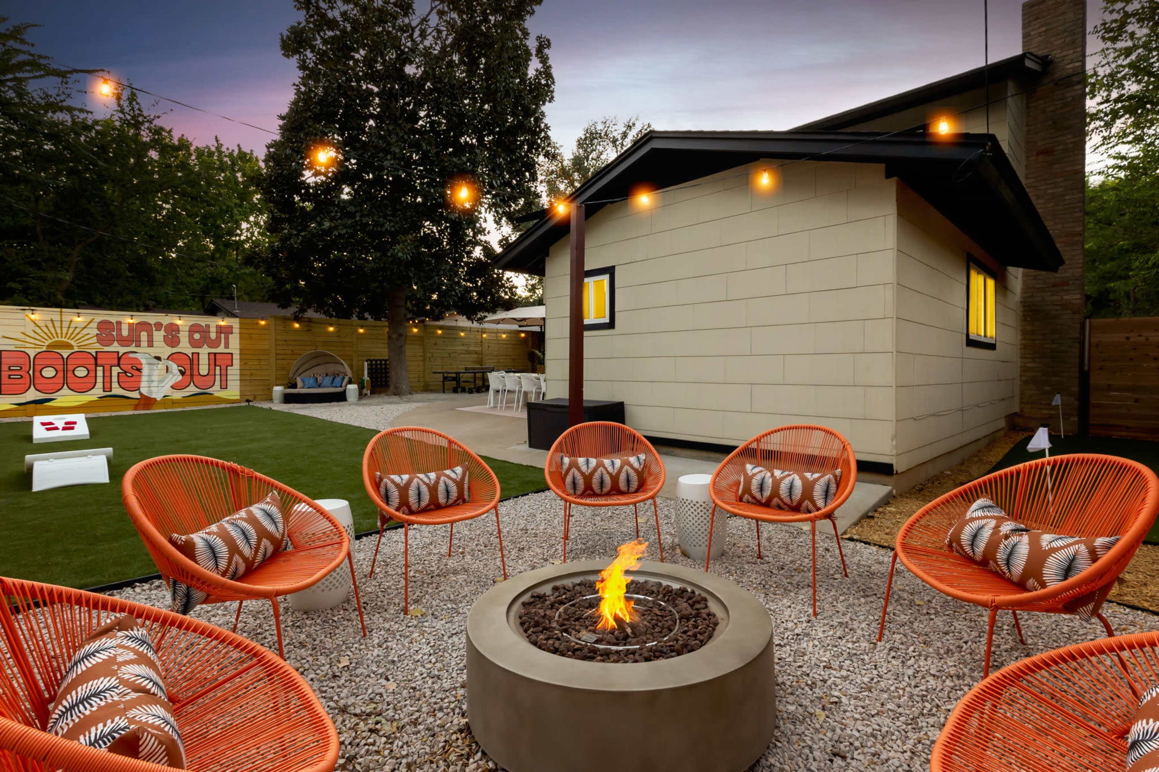 The image depicts a cozy outdoor seating area with six orange chairs arranged around a circular fire pit in a backyard setting, illuminated by string lights and featuring a decorative mural on the wall.